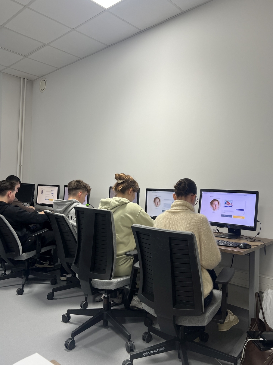 Na obrazie widać salę do pracy lub nauki z rzędem stanowisk komputerowych. Przy biurku kilka osób pracuje indywidualnie, skupiając się na ekranach, na których wyświetlane są różne strony internetowe. Pomieszczenie ma jasne, neutralne kolory, sufit podwieszany z oświetleniem oraz uporządkowaną, funkcjonalną przestrzeń do pracy.