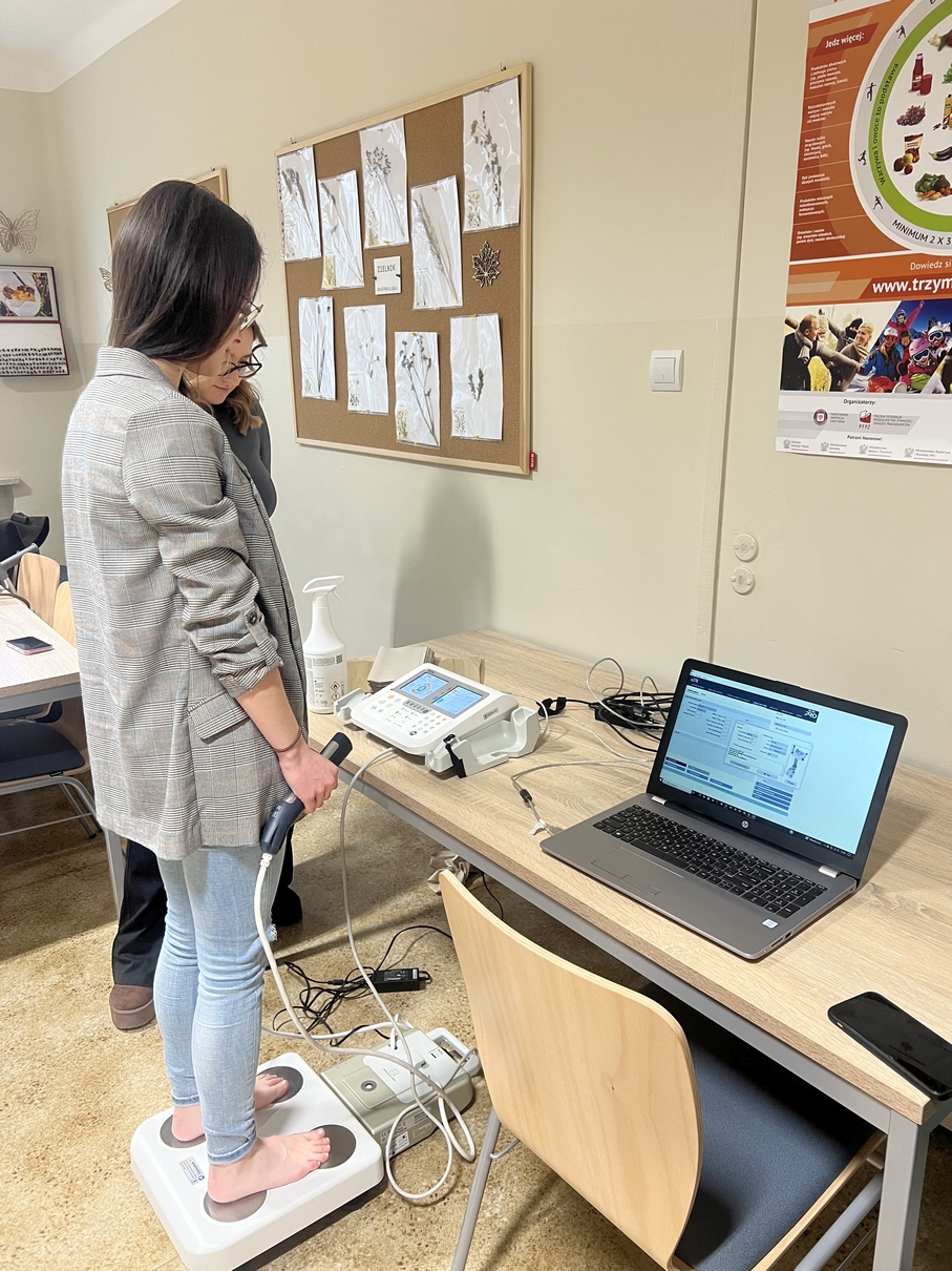 Na obrazie widoczna jest elektroniczna waga z uchwytem połączonym przewodem, używana do pomiarów. Urządzenie stoi na podłodze i jest podłączone do aparatu pomiarowego ustawionego na biurku obok otwartego laptopa z wyświetlonym interfejsem. W tle znajduje się tablica korkowa z planszami przedstawiającymi zasuszone rośliny oraz częściowo widoczny kolorowy plakat.