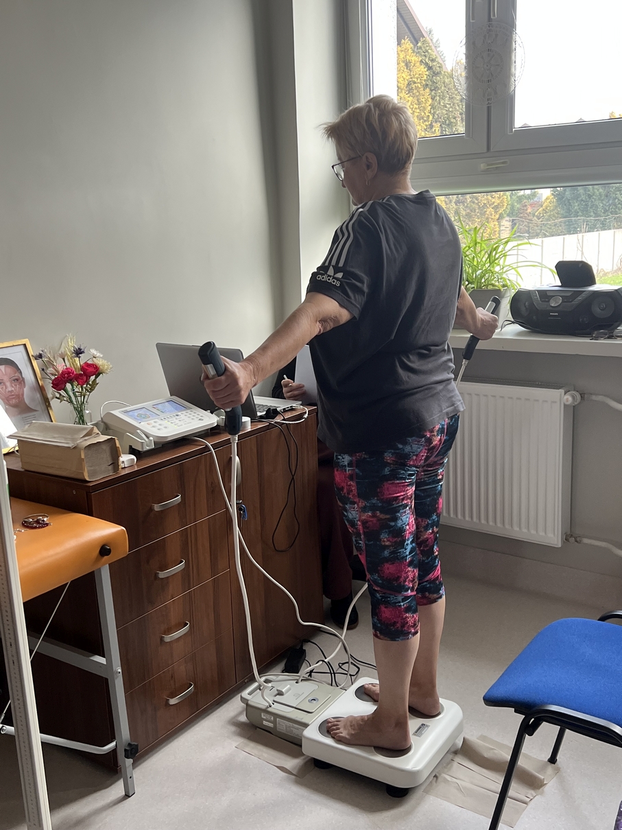 Na obrazie widoczna jest osoba stojąca na elektronicznej wadze do analizy składu ciała i trzymająca uchwyty pomiarowe. Pomiar odbywa się w jasnym pomieszczeniu z dużym oknem, przez które wpada naturalne światło i widać zieleń na zewnątrz. Wnętrze wyposażone jest w biurko z różnymi przedmiotami, krzesło oraz sprzęt elektroniczny, co nadaje mu funkcjonalny, gabinetowy charakter.