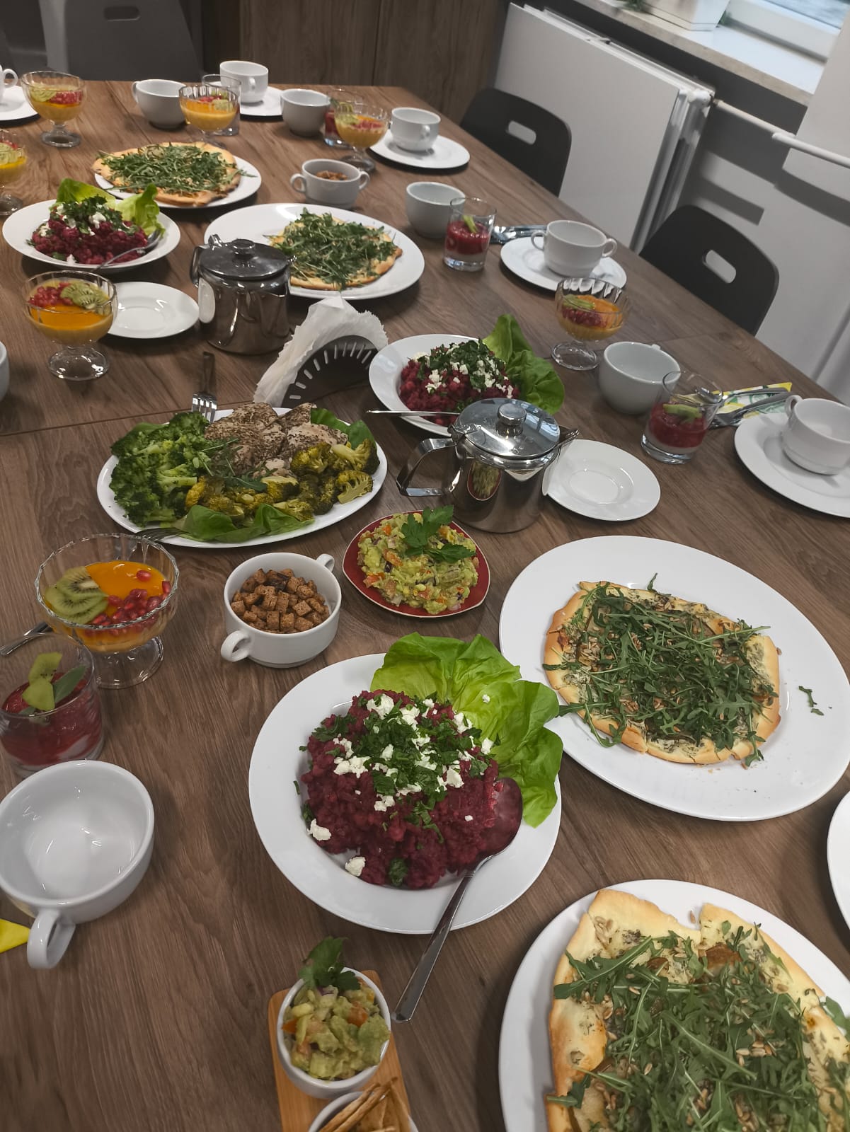 Na obrazie widać duży drewniany stół zastawiony różnorodnymi potrawami i naczyniami. Na talerzach i w misach znajdują się sałatki, risotto, pieczywo z dodatkami oraz desery w szklankach z owocami. Na stole stoją także filiżanki, spodki i czajniczki. Całość wygląda na przygotowaną do wspólnego posiłku w jasnym, nowoczesnym wnętrzu z krzesłami ustawionymi wokół stołu.