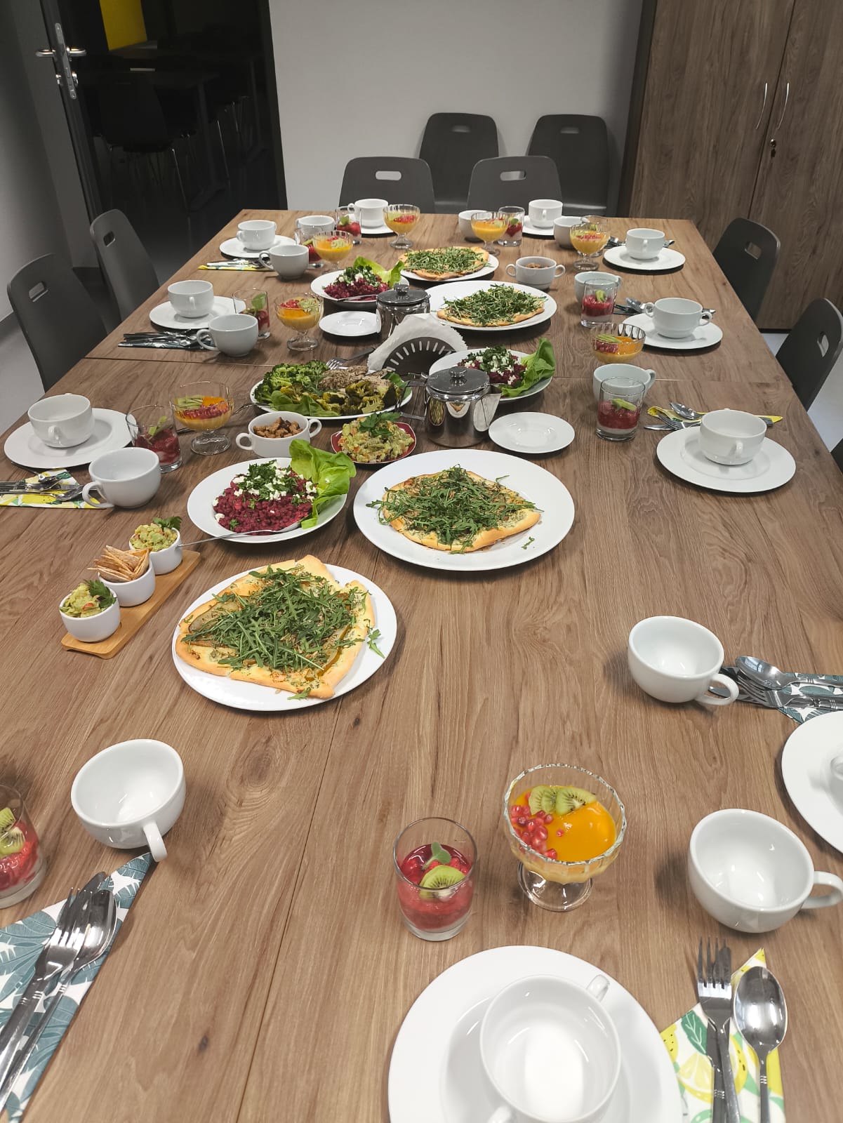 Na obrazie widać duży drewniany stół przygotowany do posiłku, otoczony krzesłami. Na stole ustawiono różne dania, w tym sałatki, potrawy z warzywami oraz desery w szklankach z owocami. Obok znajdują się filiżanki, spodki i sztućce. W tle widać minimalistyczne wnętrze w stonowanych kolorach z zamkniętymi szafkami.