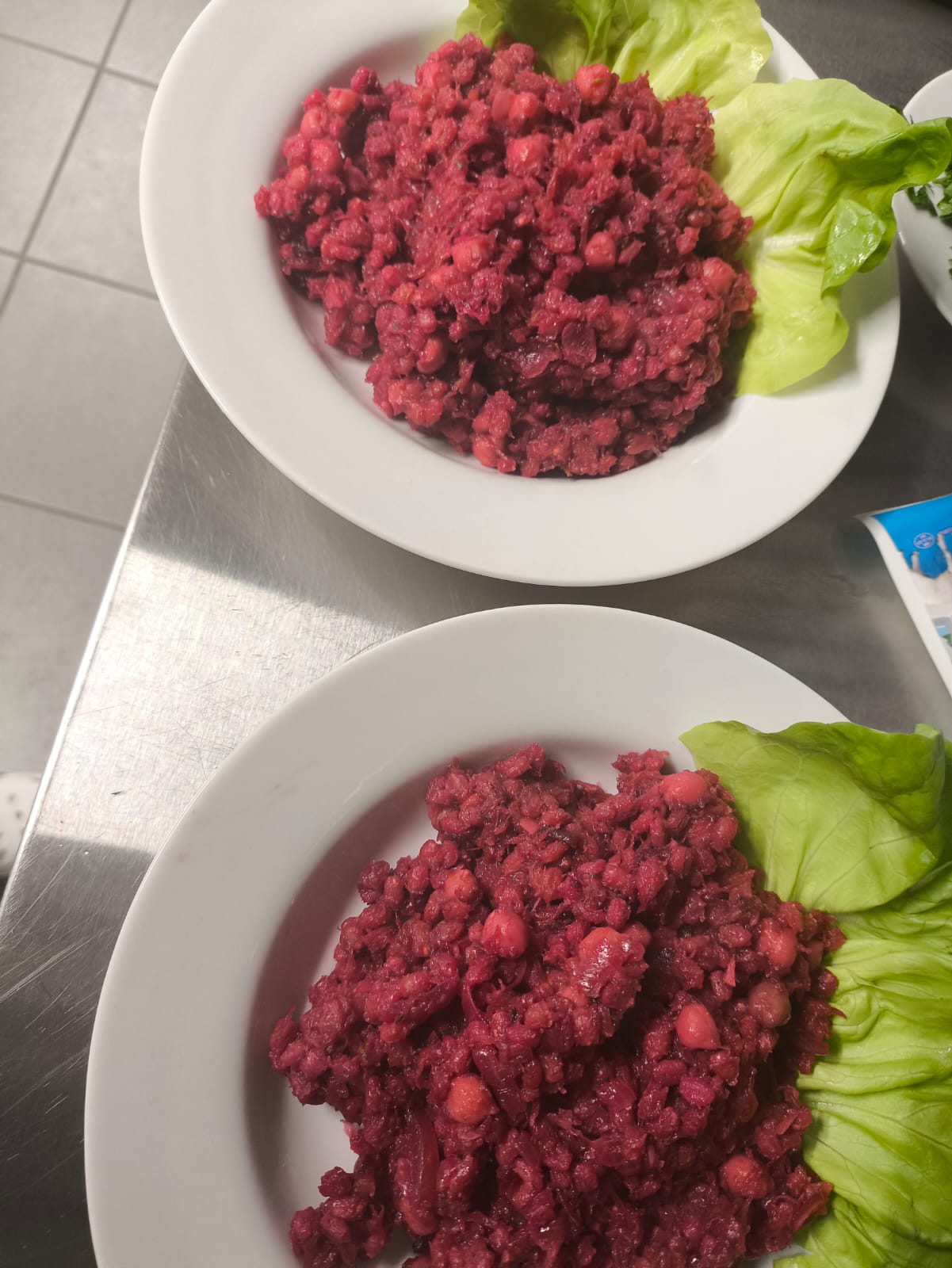 Na obrazie widać dwa talerze ustawione na metalowej powierzchni. Na każdym znajduje się porcja czerwono-fioletowej potrawy z widocznymi kawałkami warzyw i ciecierzycy. Danie udekorowane jest świeżymi liśćmi sałaty. W tle widać fragment podłogi, co sugeruje, że zdjęcie wykonano w kuchni lub jadalni.