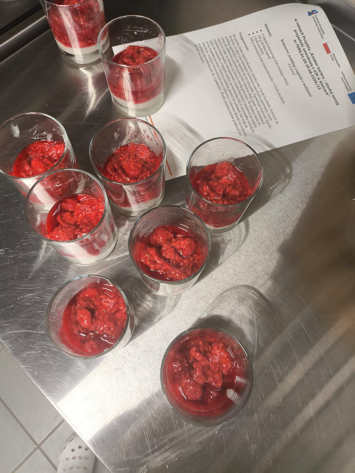 Na obrazie widać dziewięć małych szklanych pucharków ustawionych na metalowej powierzchni. W każdym znajduje się deser z jasną warstwą bazową i czerwonym owocowym sosem na wierzchu. Pucharki są rozmieszczone luźno na stole. W prawym górnym rogu leży kartka z tekstem i logo, częściowo zasłaniająca blat. Sceneria sugeruje kuchnię.