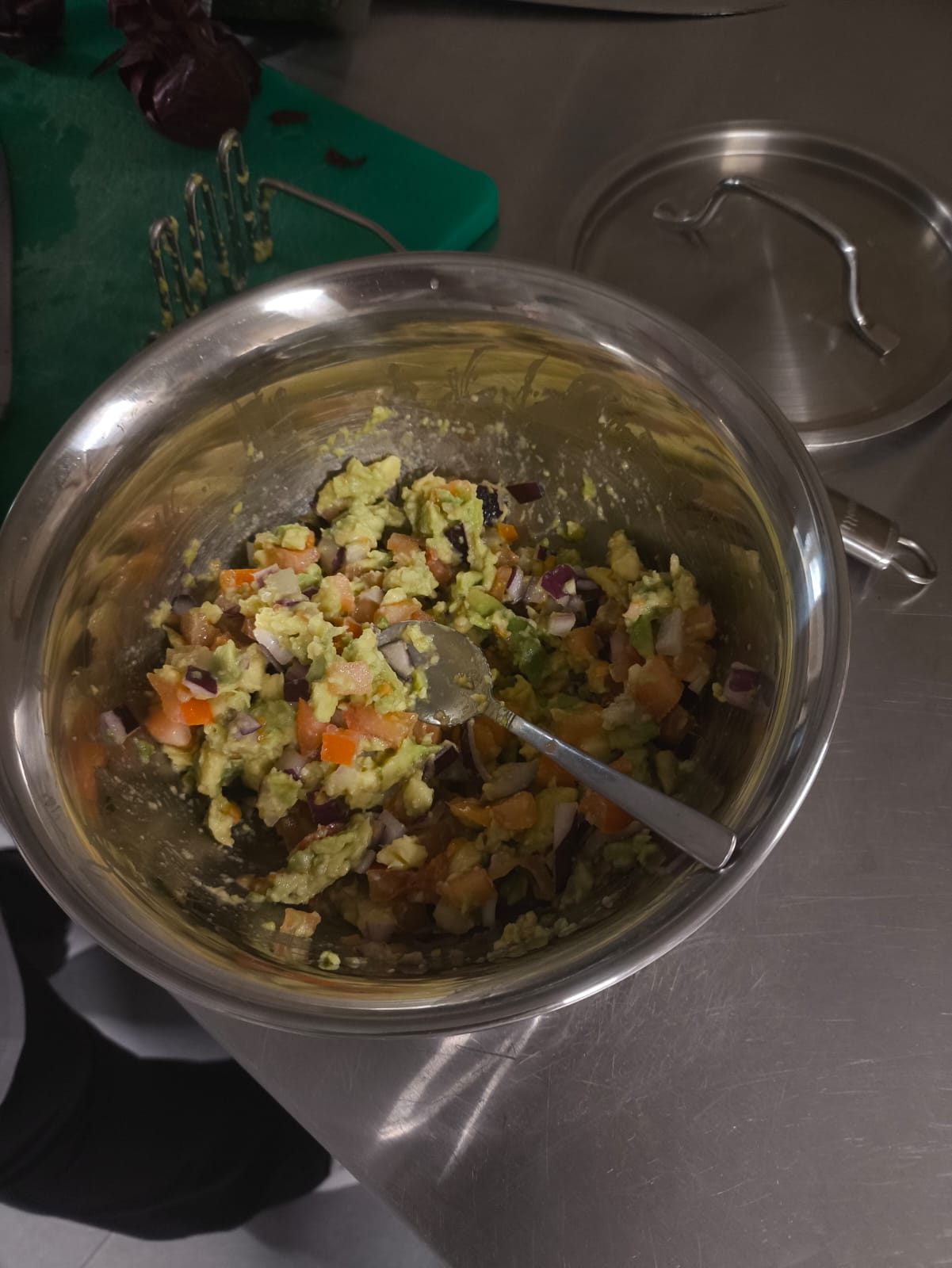 Na obrazie widać metalową miskę na metalowym blacie, wypełnioną mieszanką warzyw i rozgniecionym awokado, prawdopodobnie do sałatki lub guacamole. Obok widać zieloną deskę do krojenia z warzywami, przybór kuchenny i pokrywkę od garnka.