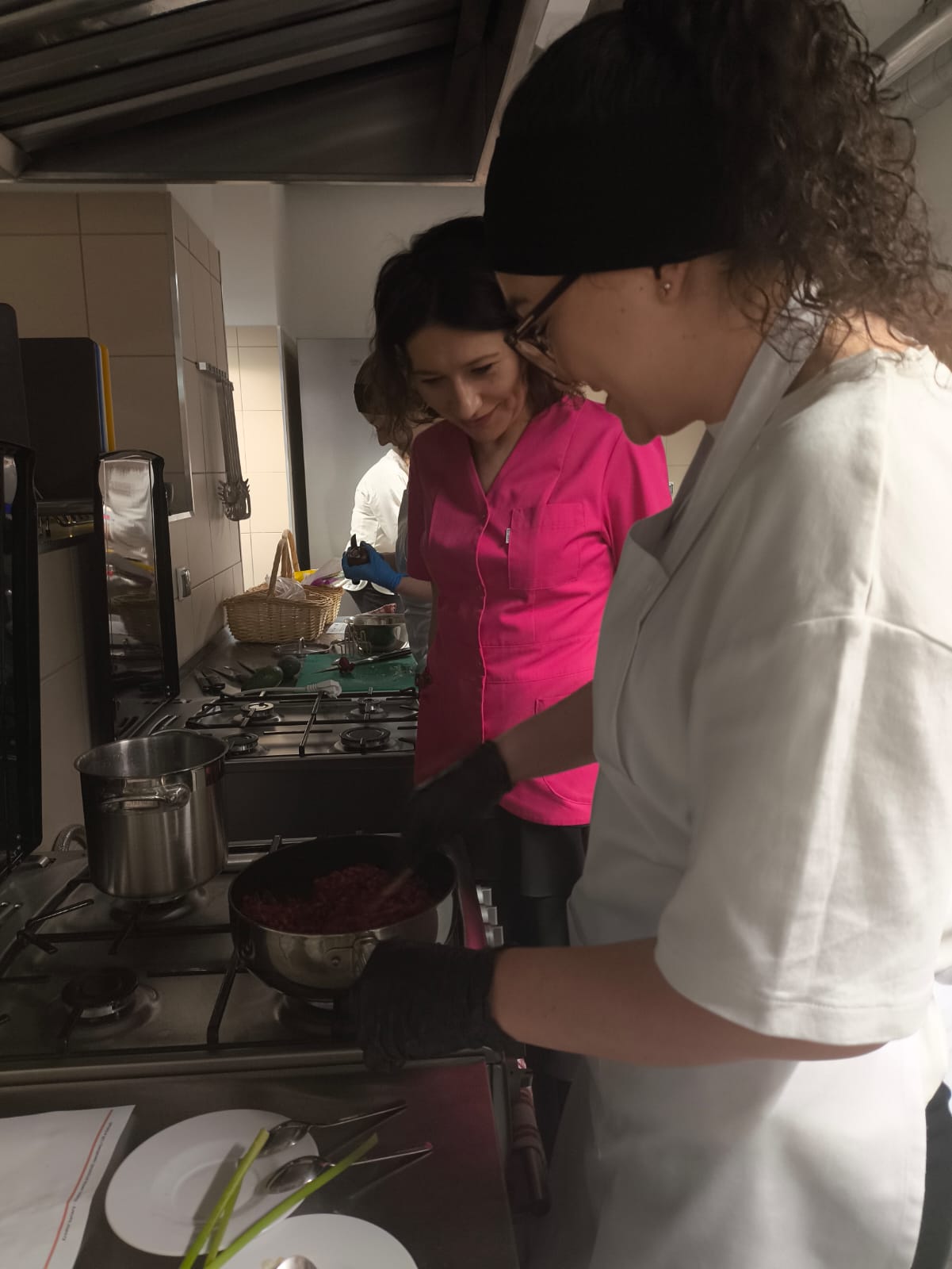 Na obrazie widać kuchnię, w której osoby przygotowują jedzenie przy metalowym blacie. Na blacie stoją garnki, przybory, talerz oraz kilka zielonych warzyw. Scena przedstawia aktywność związaną z gotowaniem.