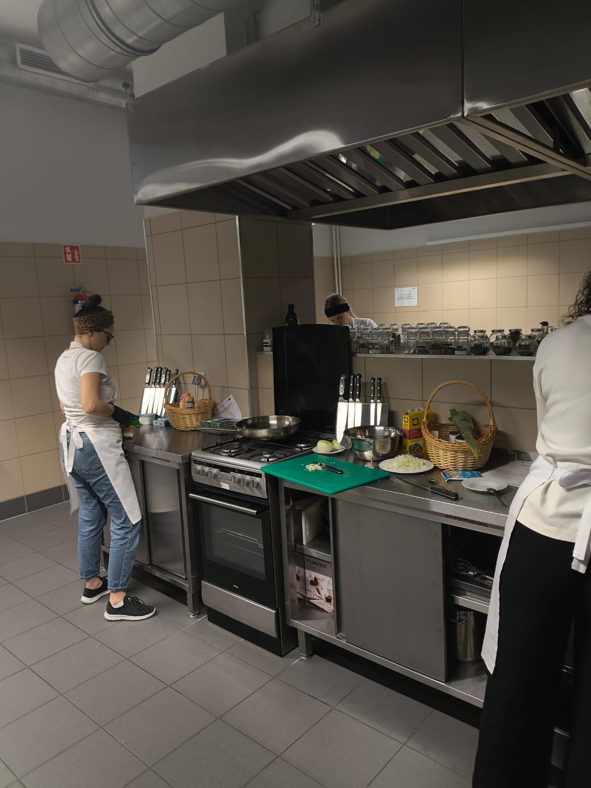 Na obrazie widać kuchnię, w której dwie osoby przygotowują jedzenie przy metalowym blacie. Na blacie leżą różne przybory i koszyki, a na kuchence stoi patelnia. W tle widoczna jest półka z słoikami, duży okap oraz kafelki na ścianach i podłodze.