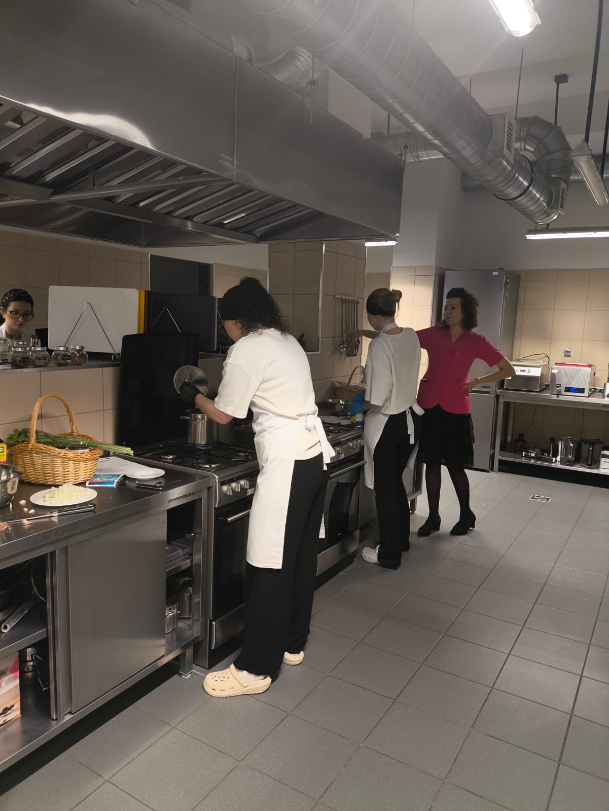 Na obrazie widać profesjonalną kuchnię z trzema osobami przygotowującymi potrawy przy metalowych blatach i kuchenkach. Na blatach leżą koszyk z warzywami, posiekana cebula i inne przybory. Wnętrze jest jasne, z widocznymi przewodami wentylacyjnymi, oświetleniem fluorescencyjnym oraz czystymi, kafelkowanymi powierzchniami.