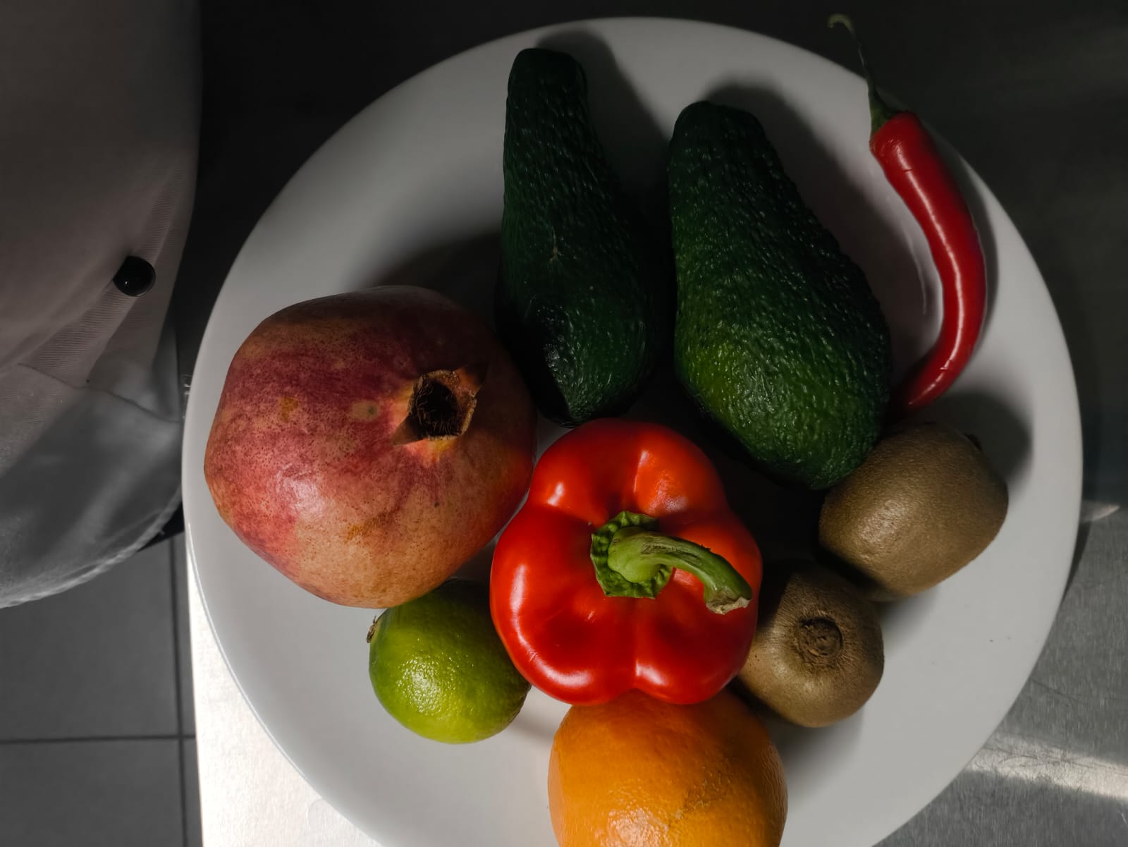 Na obrazie widać biały talerz wypełniony różnymi owocami i warzywami: papryką, granatem, limonką, pomarańczą, awokado, kiwi i chili. Tło jest ciemne, a na brzegu widać fragment ręki w białym ubraniu.