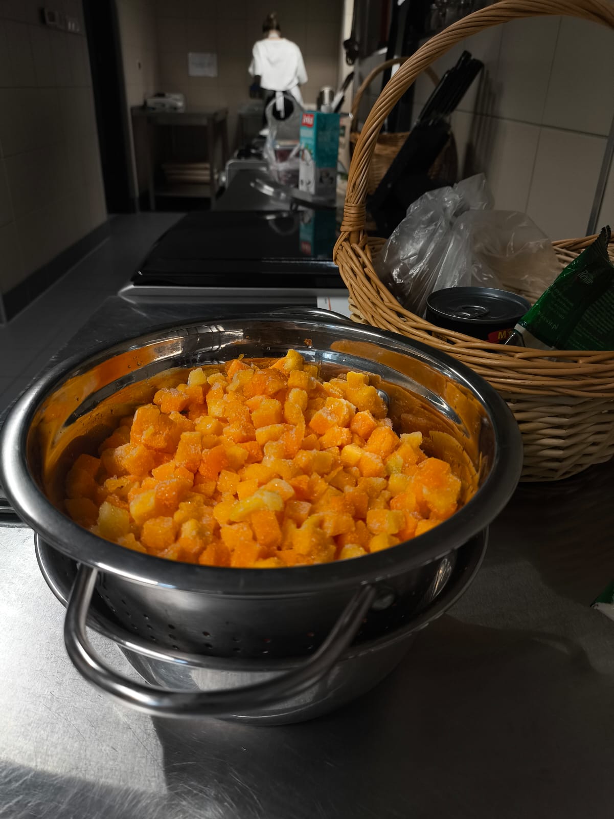 Na obrazie widać kuchnię. Na pierwszym planie znajduje się metalowy durszlak wypełniony pokrojonymi w kostkę warzywami, ustawiony na innym pojemniku. Obok stoi koszyk z różnymi produktami. W tle widoczna jest osoba pracująca przy blacie. Pomieszczenie wygląda na profesjonalną, czystą i uporządkowaną kuchnię.