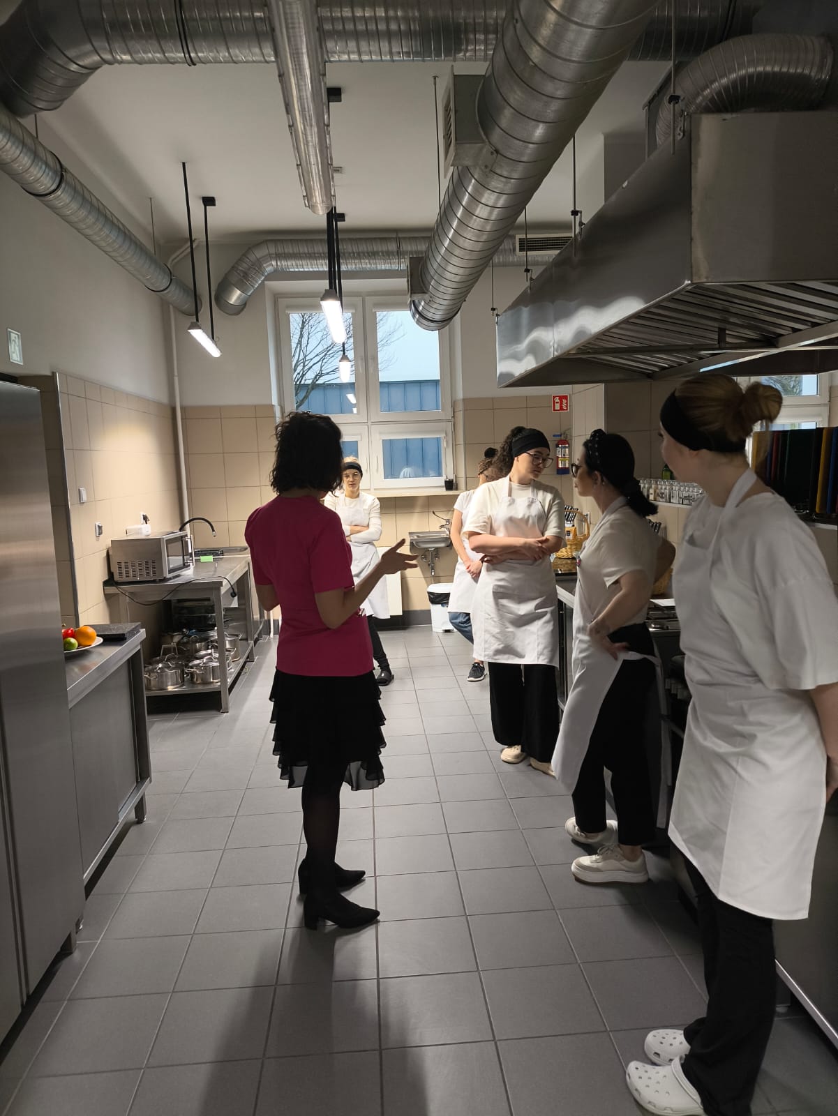 Na obrazie widać wnętrze profesjonalnej kuchni. W pomieszczeniu znajdują się metalowe blaty i półki, a podłoga jest wyłożona ciemnymi płytkami. Grupa osób stoi razem i rozmawia, prawdopodobnie podczas instruktażu. W tle widać okna wpuszczające naturalne światło.