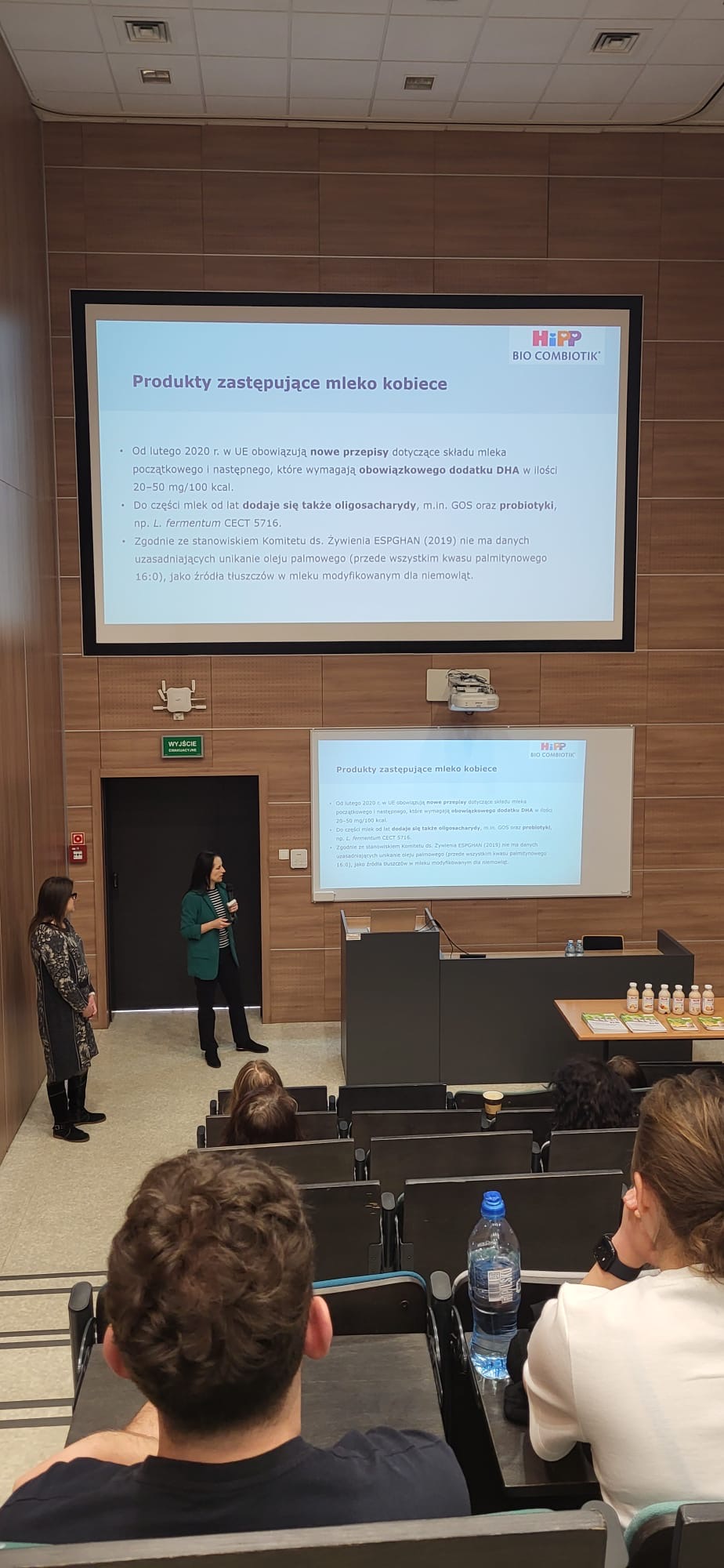 Sala wykładowa z uczestnikami siedzącymi w rzędach i obserwującymi prezentację. Z przodu sali dwie osoby stoją przy ekranach z wyświetlonym slajdem zatytułowanym „Produkty zastępujące mleko kobiece”, a jedna z nich prowadzi wystąpienie z mikrofonem. Wnętrze ma drewniane panele na ścianach, a obok stołu ustawione są materiały informacyjne oraz produkty prezentowane podczas spotkania.