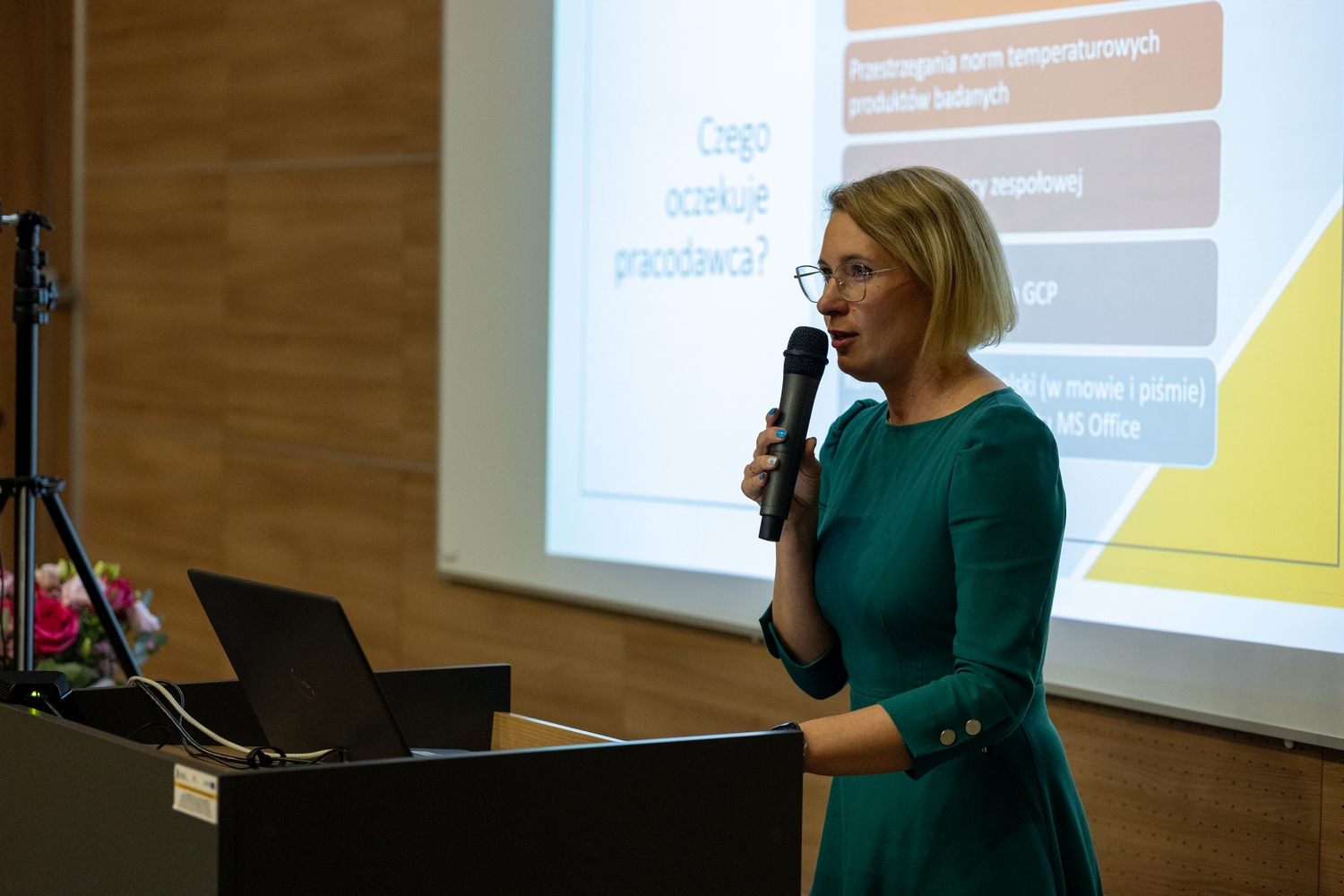 Osoba przemawia przy mównicy w sali wykładowej, trzymając mikrofon. Obok znajduje się ekran z wyświetlonym slajdem prezentacji w języku polskim oraz laptop na pulpicie. Wnętrze ma drewniane panele na ścianach, a obok widoczny jest bukiet kwiatów.