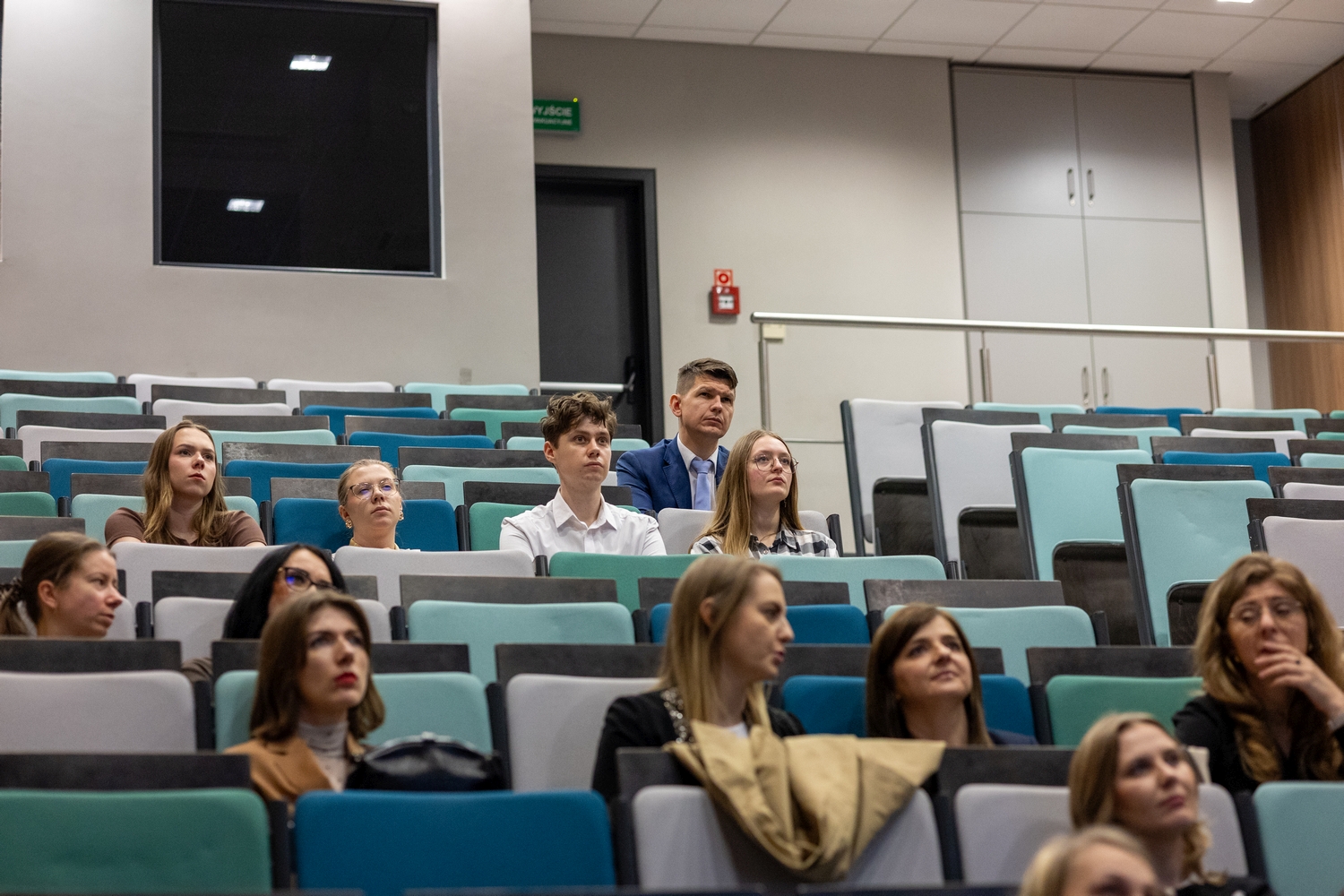 Uczestnicy siedzą w rzędach sali wykładowej z kolorowymi siedzeniami w odcieniach niebieskiego, turkusu i szarości. Publiczność obserwuje prezentację odbywającą się z przodu sali. W tle widoczne są drzwi z zielonym znakiem wyjścia ewakuacyjnego, szafki ścienne oraz jasne ściany i sufit z panelami.