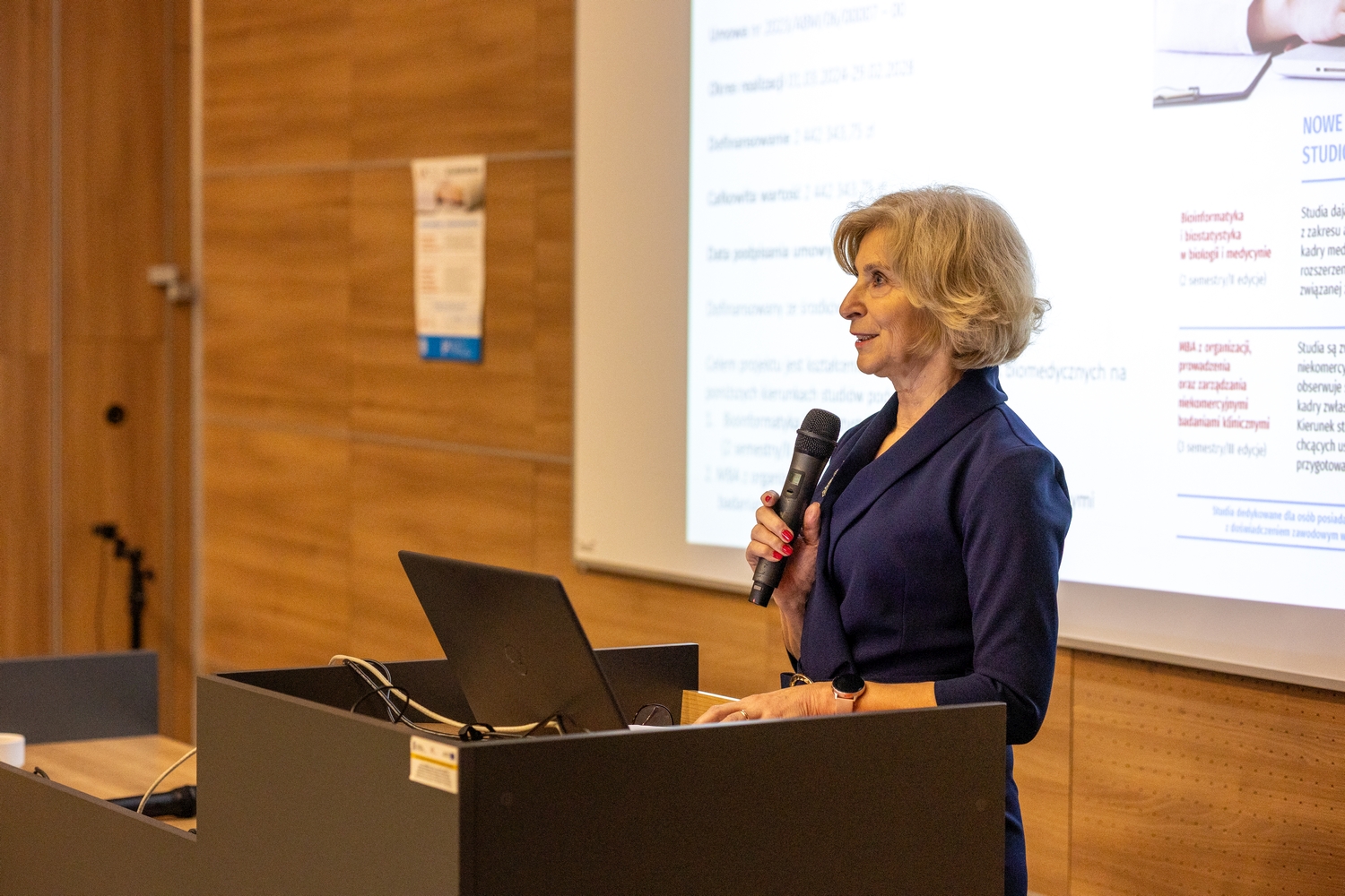 Prelegentka przemawia przy mównicy do mikrofonu, obok znajduje się laptop. W tle widoczny ekran prezentacji oraz sala konferencyjna z drewnianymi panelami i plakatem na ścianie.