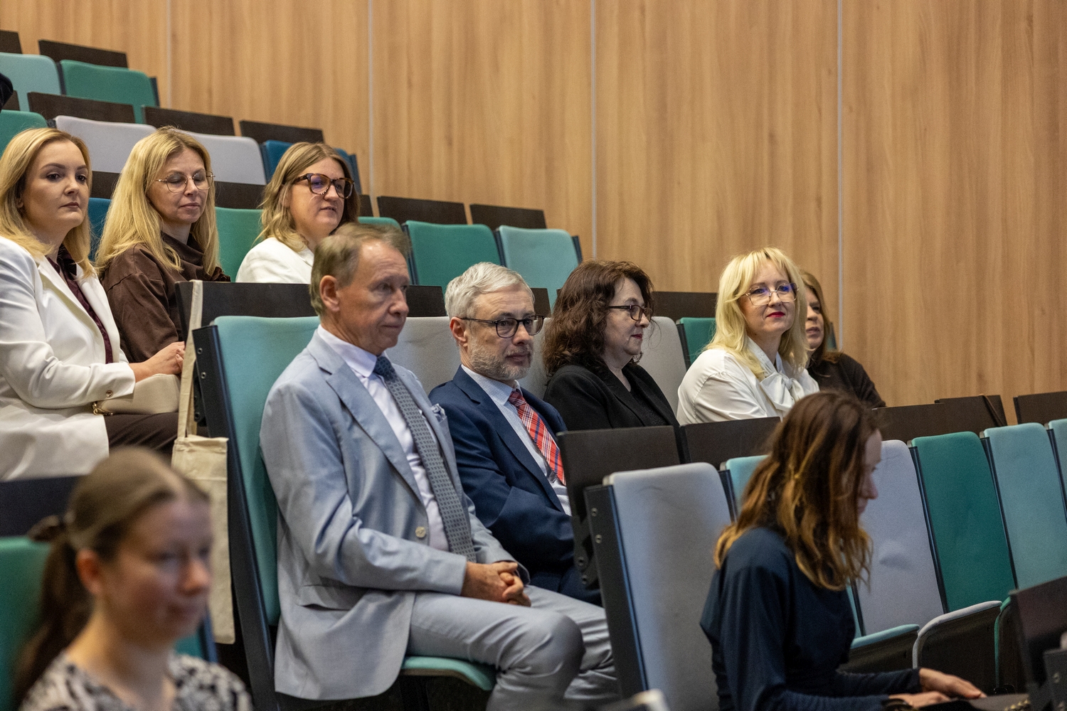 Sala wykładowa z uczestnikami siedzącymi w kilku rzędach. Wnętrze ma ściany z drewnianymi panelami. W pierwszym rzędzie siedzą uczestnicy wydarzenia, a za nimi kolejne osoby słuchające wykładu.