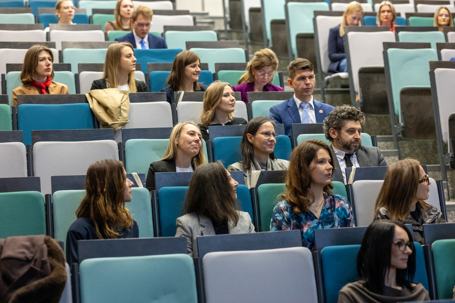 Uczestnicy siedzą w rzędach audytorium na nowoczesnych, niebiesko-szarych siedzeniach z rozkładanymi pulpitami do notowania. Osoby uczestniczą w wydarzeniu o charakterze naukowym lub profesjonalnym i słuchają prezentacji w dobrze oświetlonej sali.