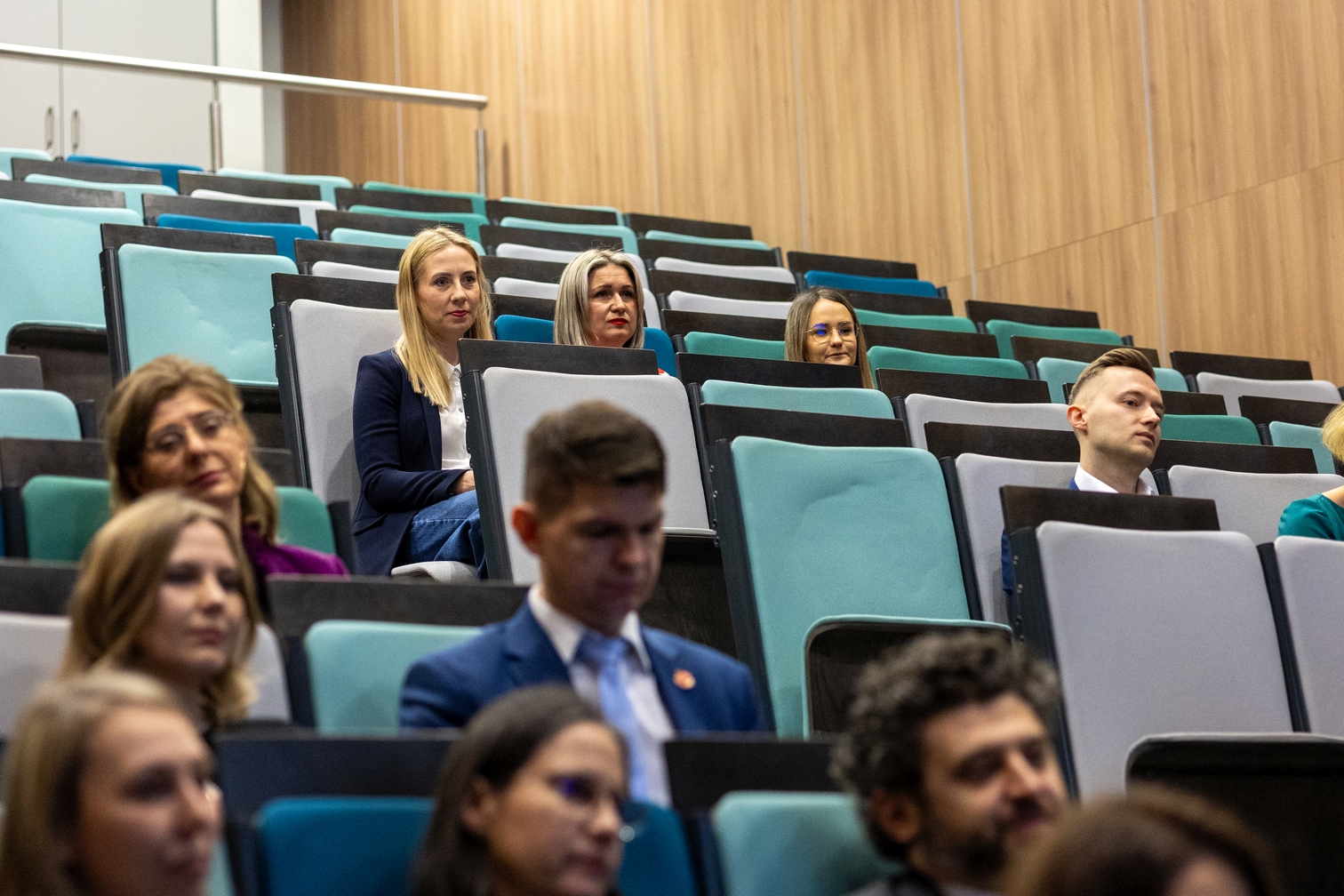 Uczestnicy siedzą w audytorium z rzędami stopniowanych siedzeń w kolorach turkusowym i szarym. Sala ma nowoczesny wystrój z jasnymi drewnianymi panelami na ścianach. Publiczność uczestniczy w wykładzie lub prezentacji i jest zwrócona w stronę sceny.