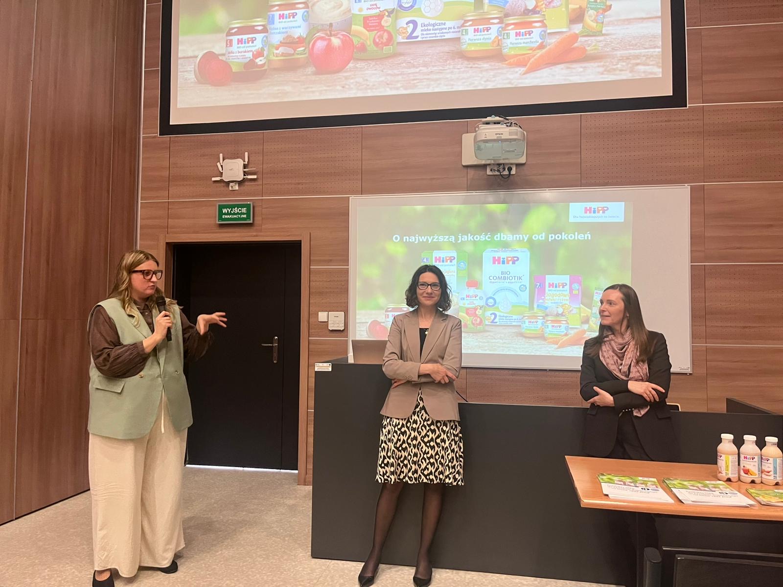 Trzy osoby stoją w sali konferencyjnej przed ekranami z wyświetloną prezentacją. Jedna z nich przemawia do mikrofonu, prowadząc spotkanie lub prezentację. W tle widoczne są drewniane panele ścienne, a na stole ustawione są materiały promocyjne związane z prezentowanym produktem.