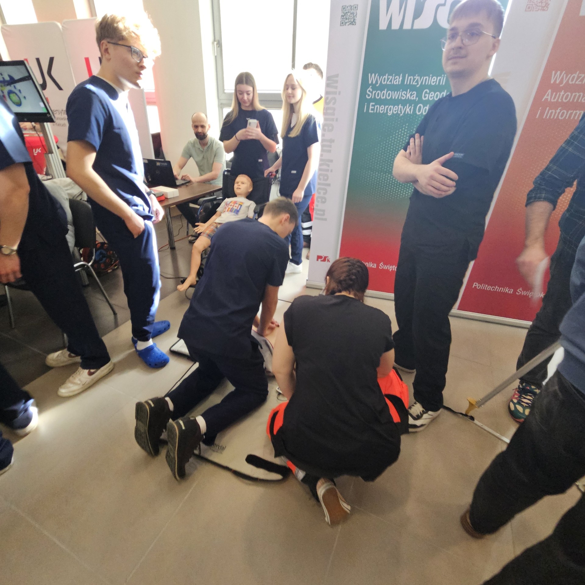 Uczestnicy biorą udział w szkoleniu z zakresu pierwszej pomocy, skupiając się wokół fantomu, na którym demonstrowane są czynności ratunkowe. Część osób obserwuje, a część aktywnie uczestniczy w ćwiczeniach. W tle widoczne są banery informacyjne oraz stanowisko z laptopem, co wskazuje na edukacyjny charakter wydarzenia.