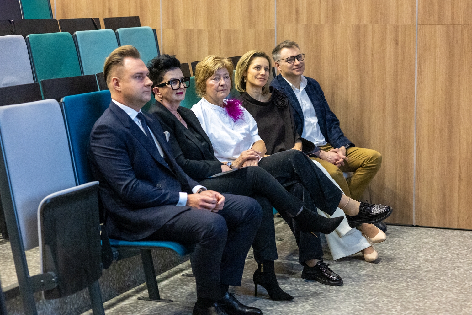 Uczestnicy siedzą w rzędzie kolorowych siedzeń w sali wykładowej. Wnętrze ma drewniane panele na ścianach oraz szarą, wzorzystą wykładzinę na podłodze. Publiczność bierze udział w wydarzeniu o charakterze naukowym lub konferencyjnym.