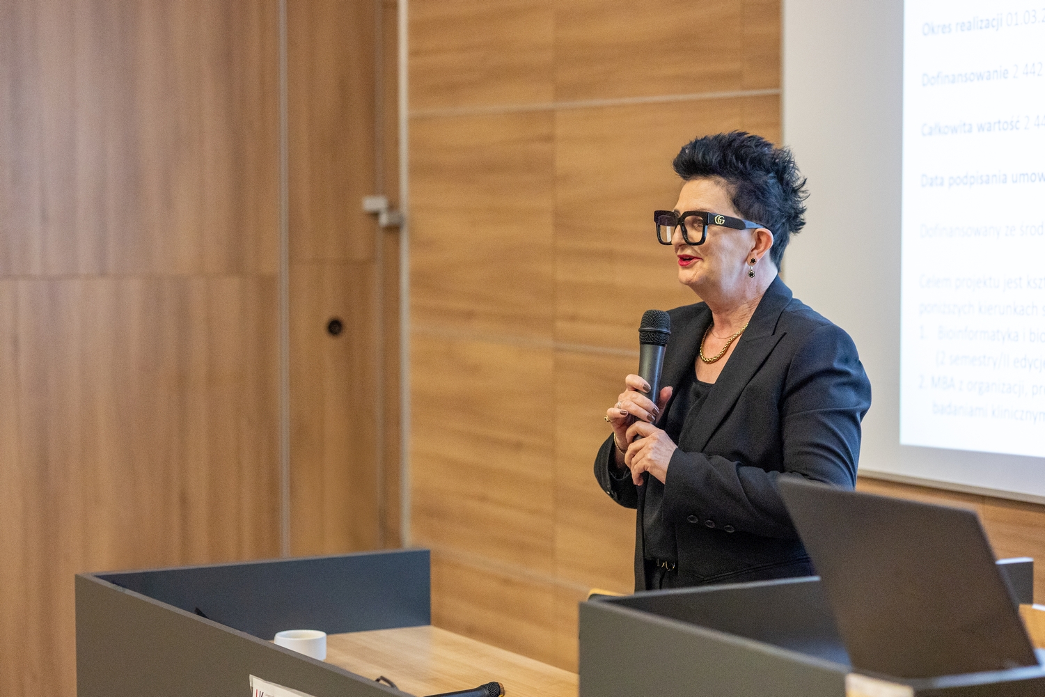 Osoba przemawia przy mównicy, trzymając mikrofon. Na pulpicie znajduje się kubek, a w tle widoczna jest ściana z drewnianych paneli oraz ekran z wyświetlaną prezentacją.