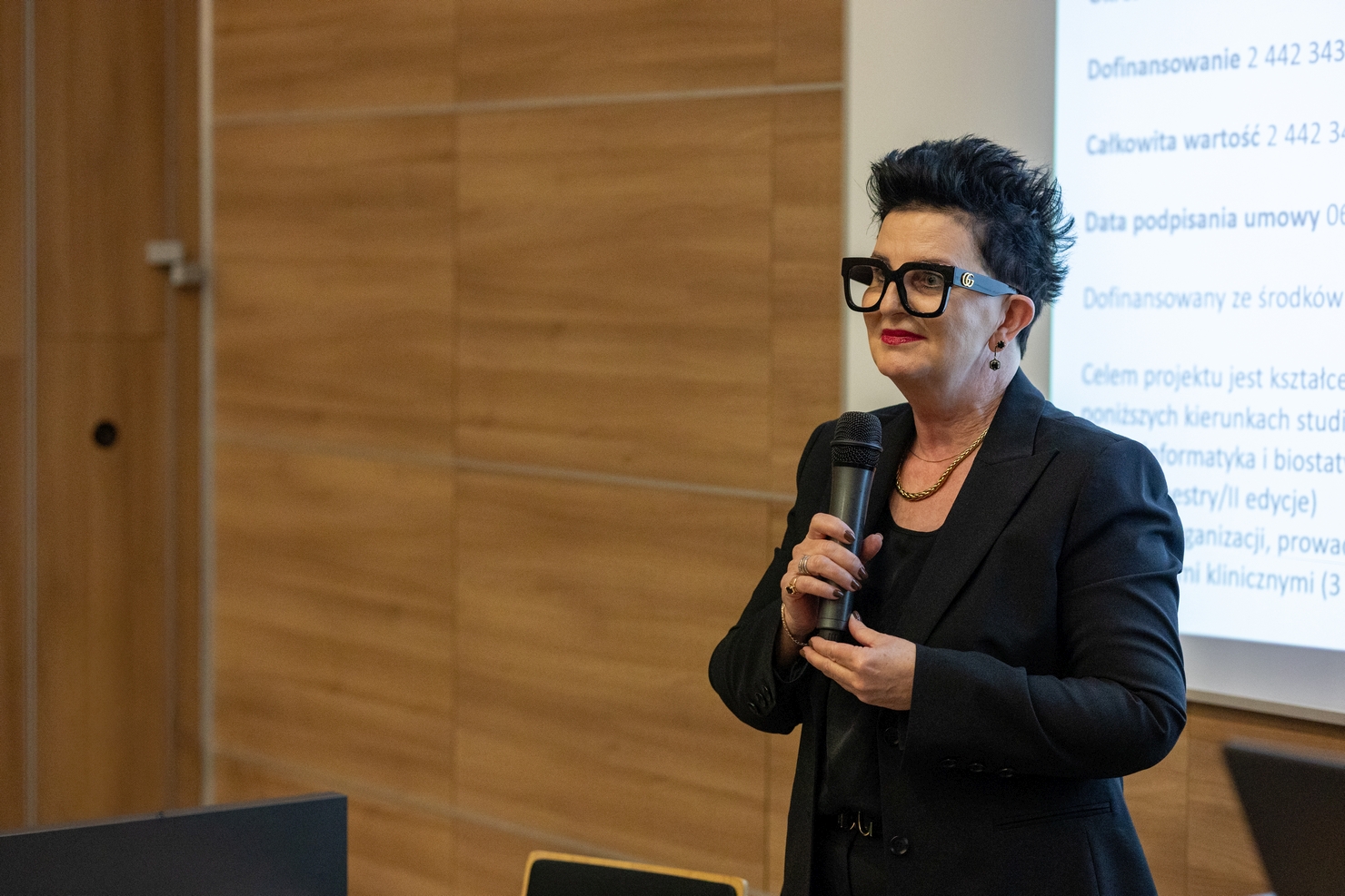 Osoba przemawia do mikrofonu podczas wystąpienia. W tle widoczna jest ściana z drewnianych paneli oraz ekran z wyświetlaną prezentacją w sali konferencyjnej lub wykładowej.