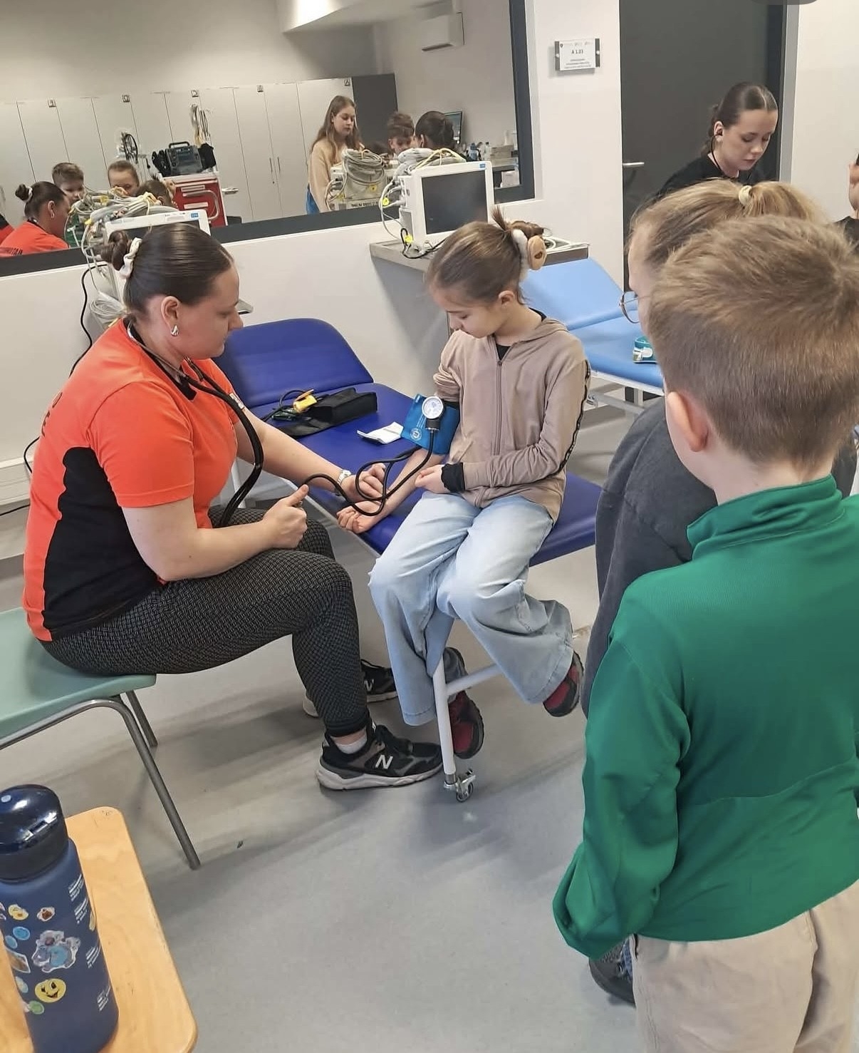 Osoba przeprowadza pomiar ciśnienia tętniczego u dziecka, demonstrując badanie w otoczeniu przypominającym salę medyczną lub edukacyjną. W tle widoczne są urządzenia medyczne, szafki oraz lustro odbijające przebieg zajęć, a inni uczestnicy obserwują pokaz.