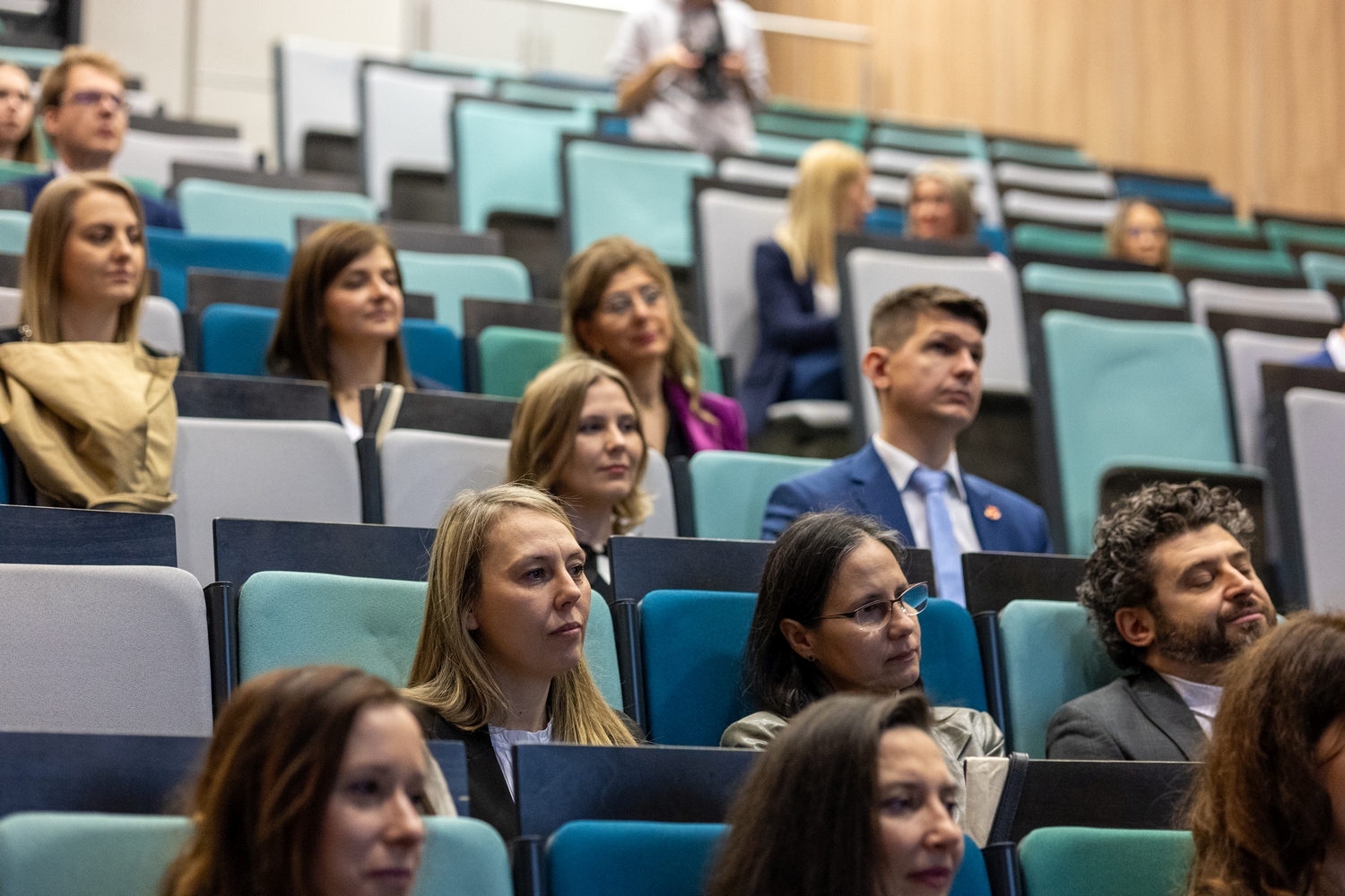 Uczestnicy siedzą w rzędach audytorium na stopniowanych siedzeniach w odcieniach niebieskiego, turkusu i szarości. Publiczność jest zwrócona w stronę sceny, obserwując prezentację, a w tle widoczne są kolejne rzędy miejsc oraz drewniane ściany sali.