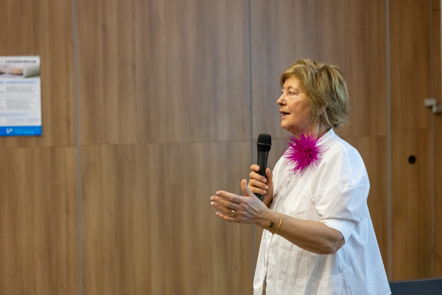 Osoba przemawia do mikrofonu podczas wystąpienia. W tle widoczna jest ściana z drewnianych paneli oraz plakat związany z wydarzeniem o charakterze informacyjnym lub edukacyjnym.