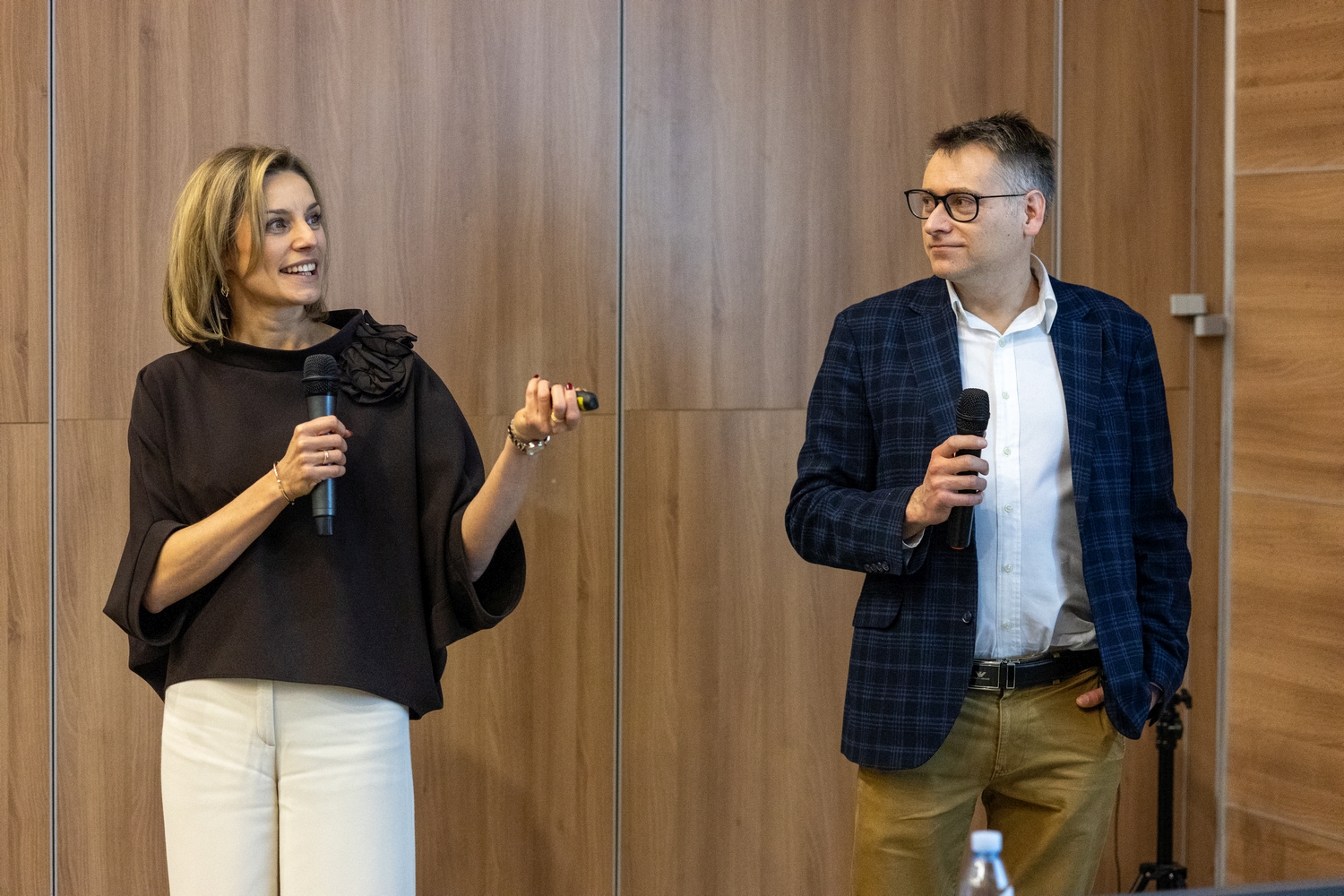 Dwie osoby stoją przed drewnianą ścianą i prowadzą wystąpienie, trzymając mikrofony podczas wydarzenia o charakterze konferencyjnym lub edukacyjnym.