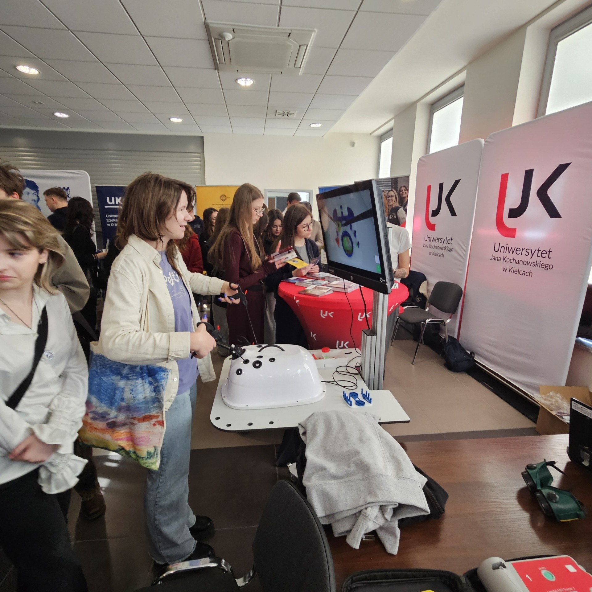 Uczestnicy biorą udział w wydarzeniu edukacyjnym lub targach, gdzie prezentowane są stanowiska demonstracyjne. Na pierwszym planie osoba korzysta z symulatora połączonego z monitorem, obsługując urządzenie za pomocą kontrolerów. W tle widoczne są stoiska z materiałami promocyjnymi oraz banery uczelni, co wskazuje na akademicki charakter wydarzenia.