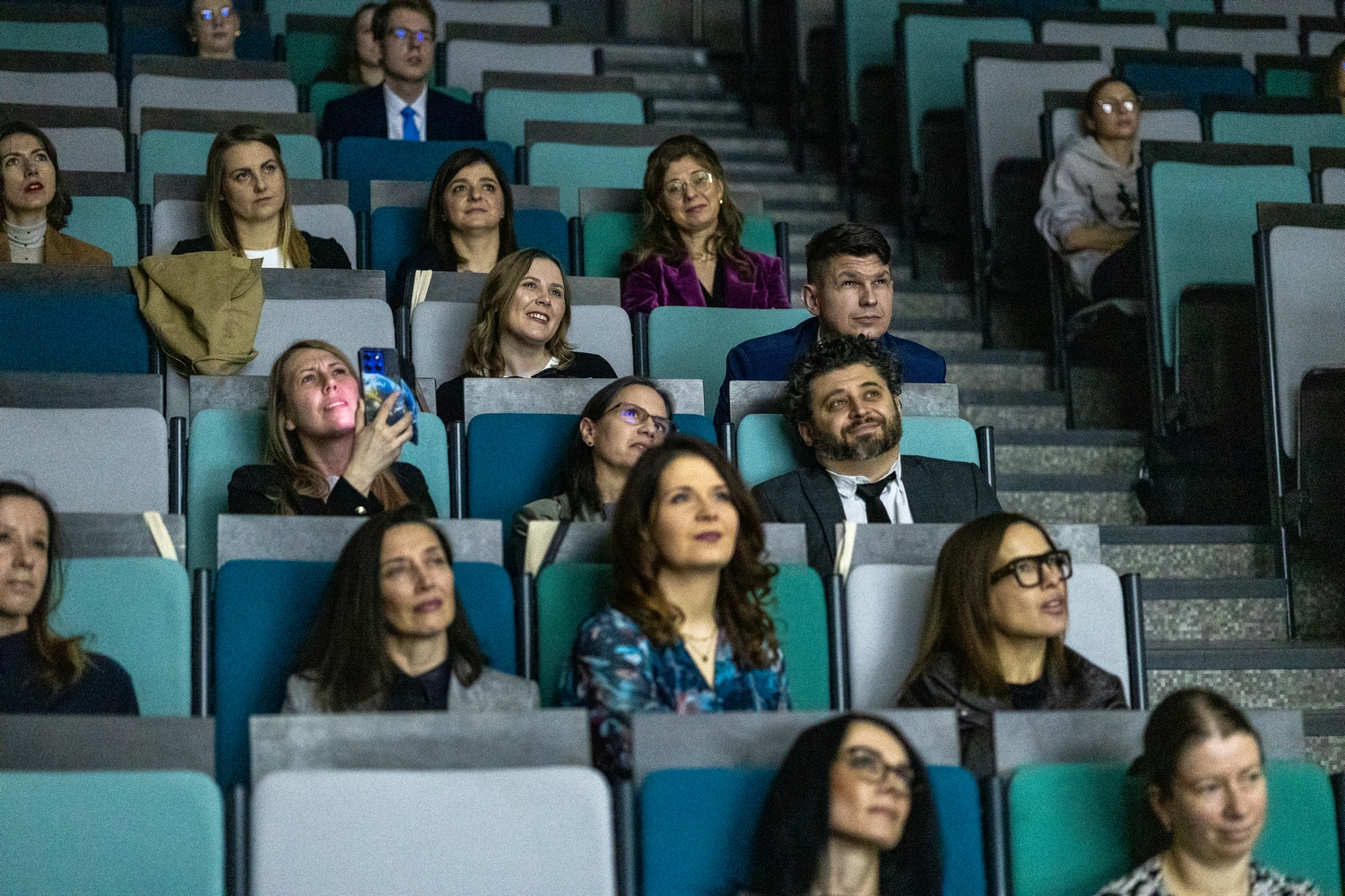 Uczestnicy siedzą w rzędach audytorium na kolorowych siedzeniach w odcieniach turkusu i szarości. Publiczność, składająca się z kobiet i mężczyzn, obserwuje prezentację odbywającą się z przodu sali. Jedna z osób trzyma telefon, prawdopodobnie wykonując zdjęcie lub nagrywając wydarzenie. Wnętrze ma nowoczesny charakter i sprzyja atmosferze skupienia podczas wykładu lub konferencji.