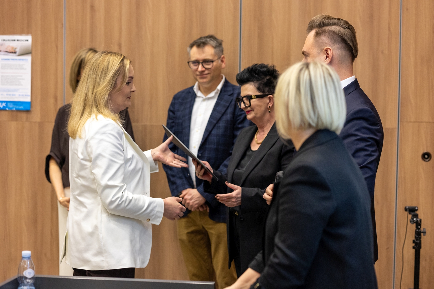 Podczas wydarzenia jedna osoba wręcza drugiej certyfikat lub dyplom. W tle stoją inni uczestnicy obserwujący uroczystość. Scena odbywa się w sali z drewnianą ścianą oraz plakatem związanym z wydarzeniem.