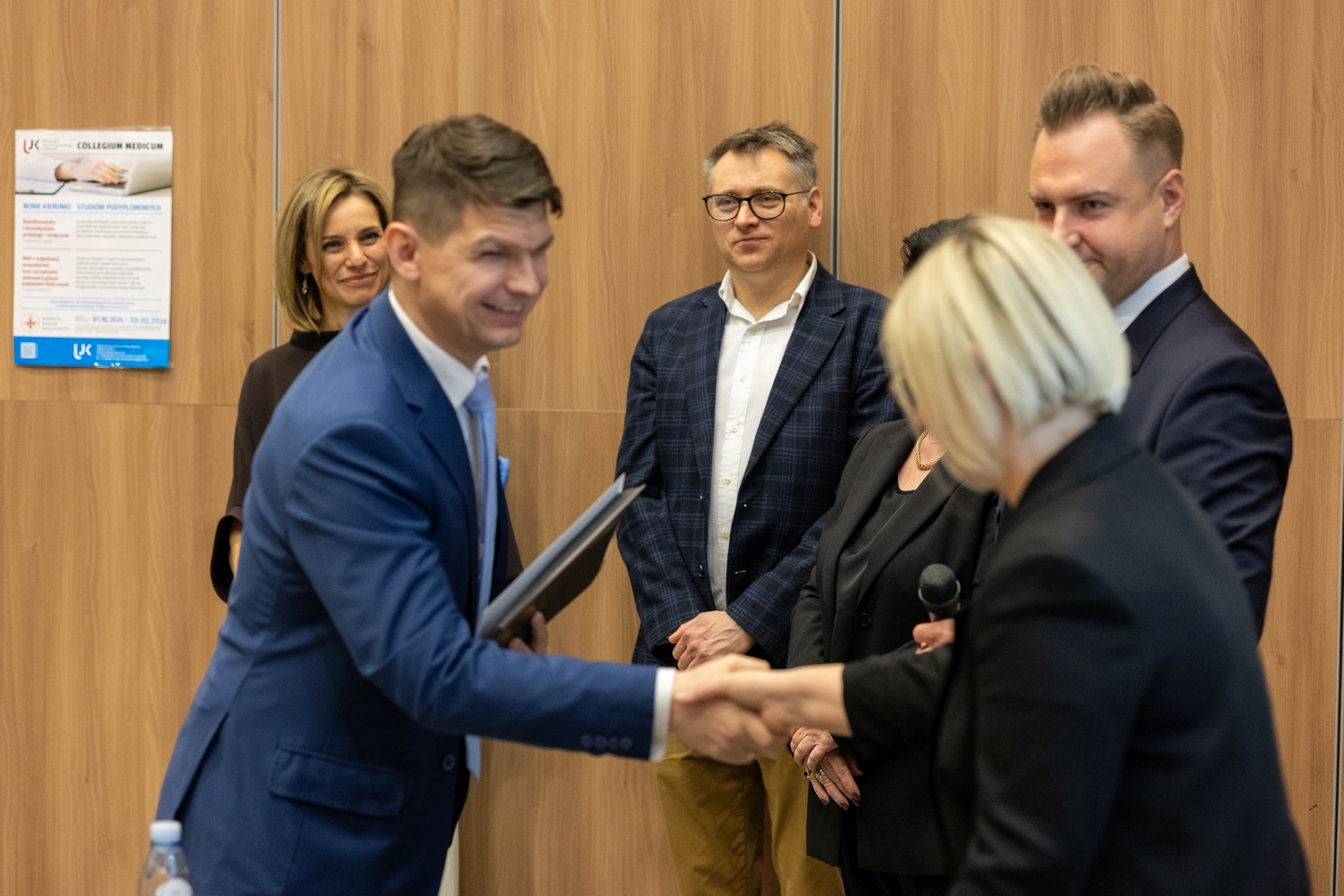 Podczas wydarzenia jedna osoba wręcza drugiej certyfikat lub dyplom. W tle stoją inni uczestnicy obserwujący uroczystość. Scena odbywa się w sali z drewnianą ścianą oraz plakatem związanym z wydarzeniem.