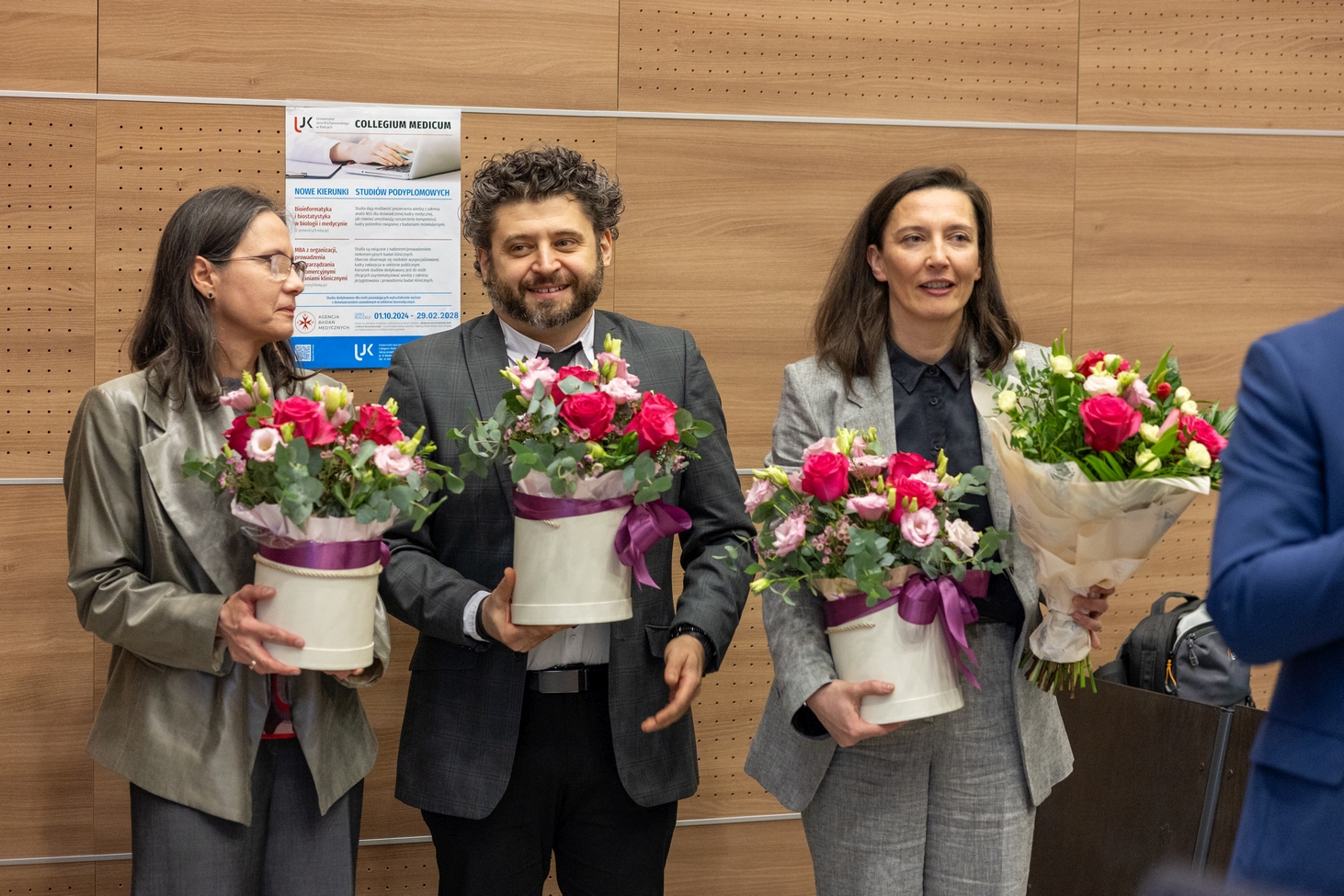 Trzy osoby stoją obok siebie w sali i trzymają bukiety kwiatów, prawdopodobnie podczas podziękowań lub zakończenia wydarzenia. W tle widoczna jest ściana z drewnianych paneli oraz plakat związany z wydarzeniem.