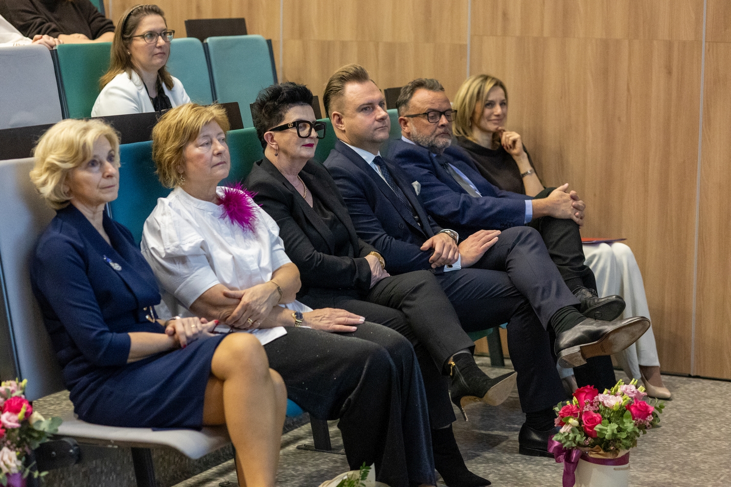 Grupa uczestników siedzi w rzędzie audytorium i obserwuje prezentację lub wystąpienie odbywające się z przodu sali. W tle widoczne są jasne drewniane panele ścienne, a przed siedzeniami ustawione są bukiety kolorowych kwiatów, co sugeruje uroczysty charakter wydarzenia.