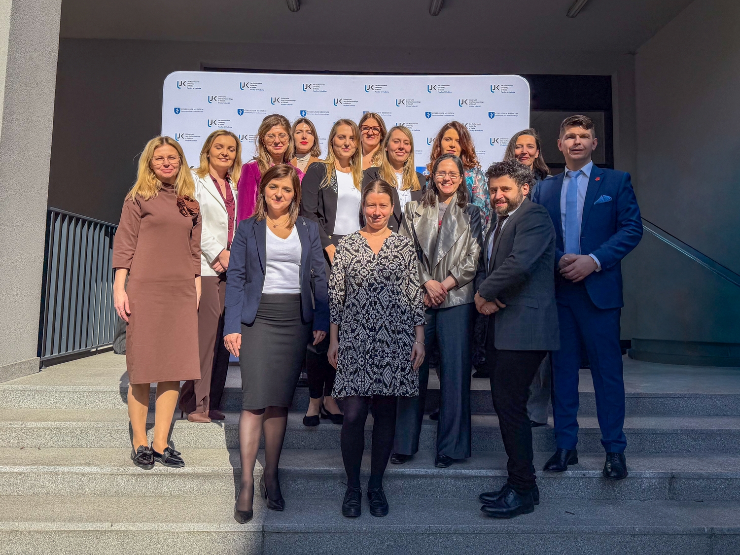 Grupa uczestników wydarzenia pozuje do wspólnego zdjęcia na schodach. W tle widoczny jest biały baner z logotypami związanymi z konferencją lub wydarzeniem. Zdjęcie wykonano na zewnątrz, przy schodach z metalową