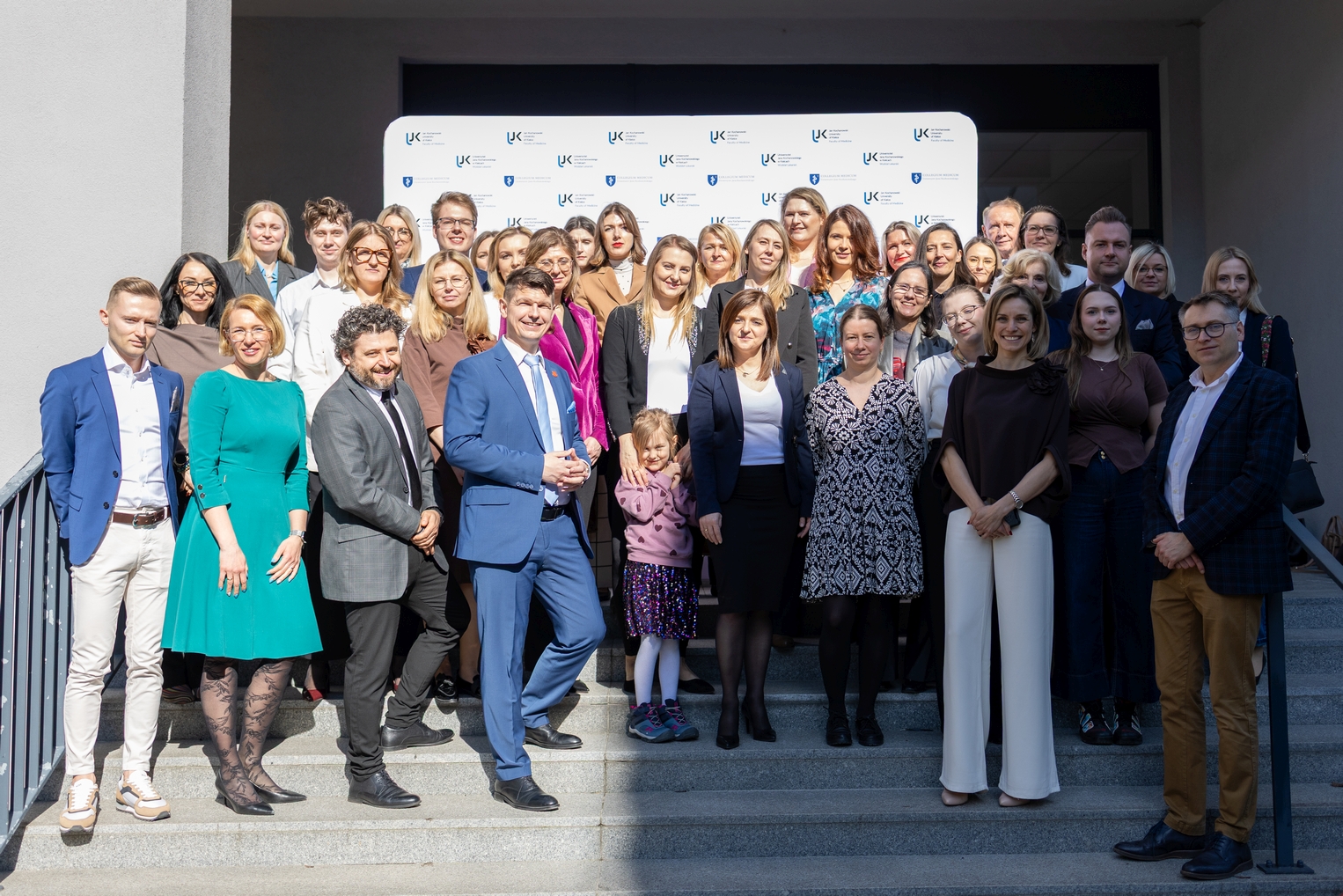 Grupa uczestników wydarzenia pozuje do wspólnego zdjęcia na schodach. W tle widoczny jest biały baner z logotypami związanymi z konferencją lub wydarzeniem. Zdjęcie wykonano na zewnątrz, przy schodach z metalową