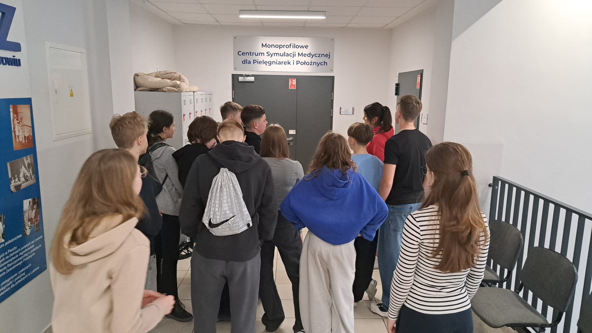 Obraz przedstawia grupę osób stojących w jasnym korytarzu z białymi ścianami i kafelkową podłogą, skierowanych w stronę szarej drzwi. Po lewej widać rząd szafek, a po prawej metalowe barierki i kilka pustych krzeseł.