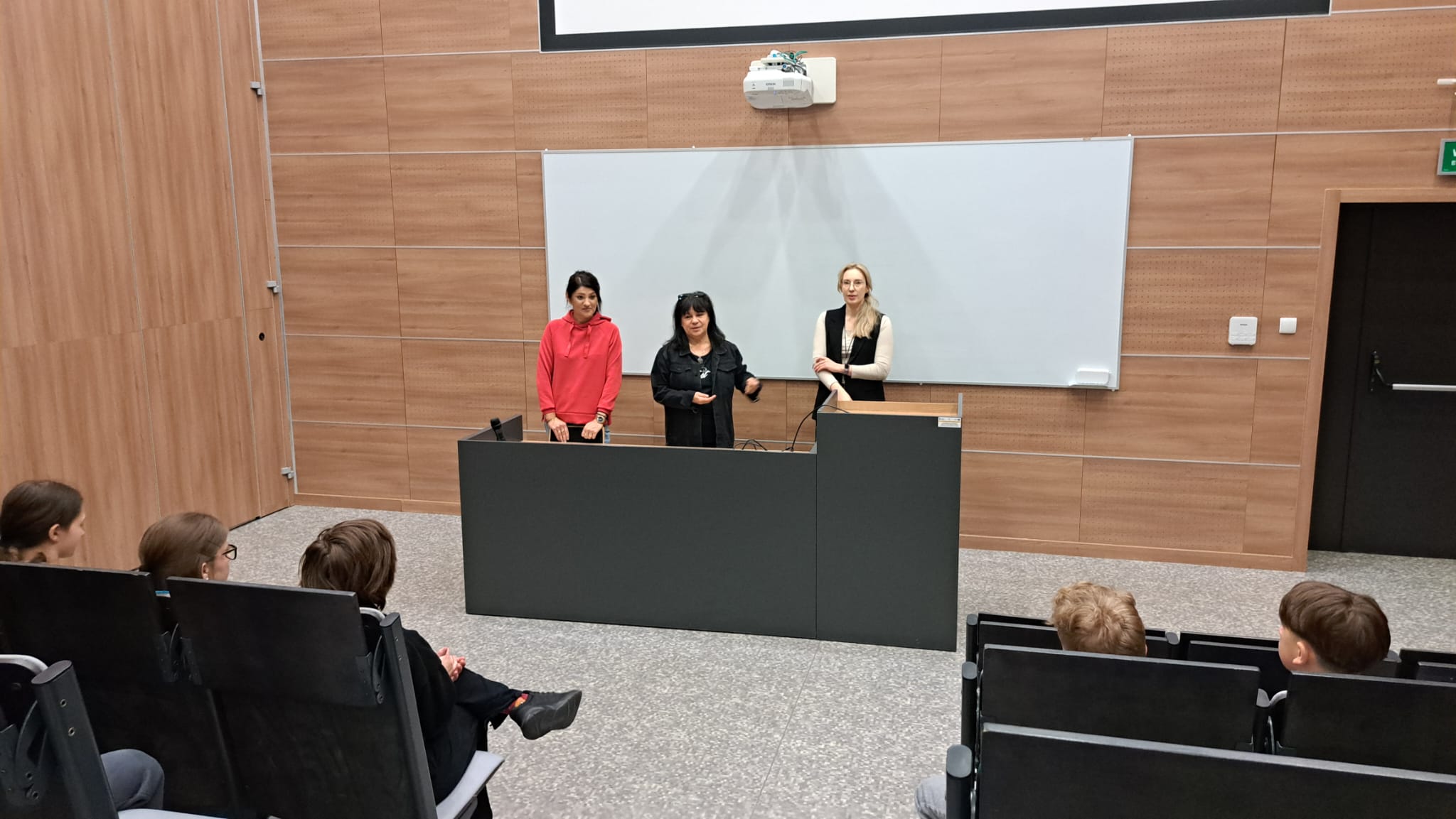 Obraz przedstawia salę wykładową z jasnymi drewnianymi panelami. Z przodu przy podium stoją osoby prowadzące prezentację, a przed nimi siedzą uczestnicy. Nad tablicą zamontowano projektor, a podłoga ma szaro-wzorzysty kolor.