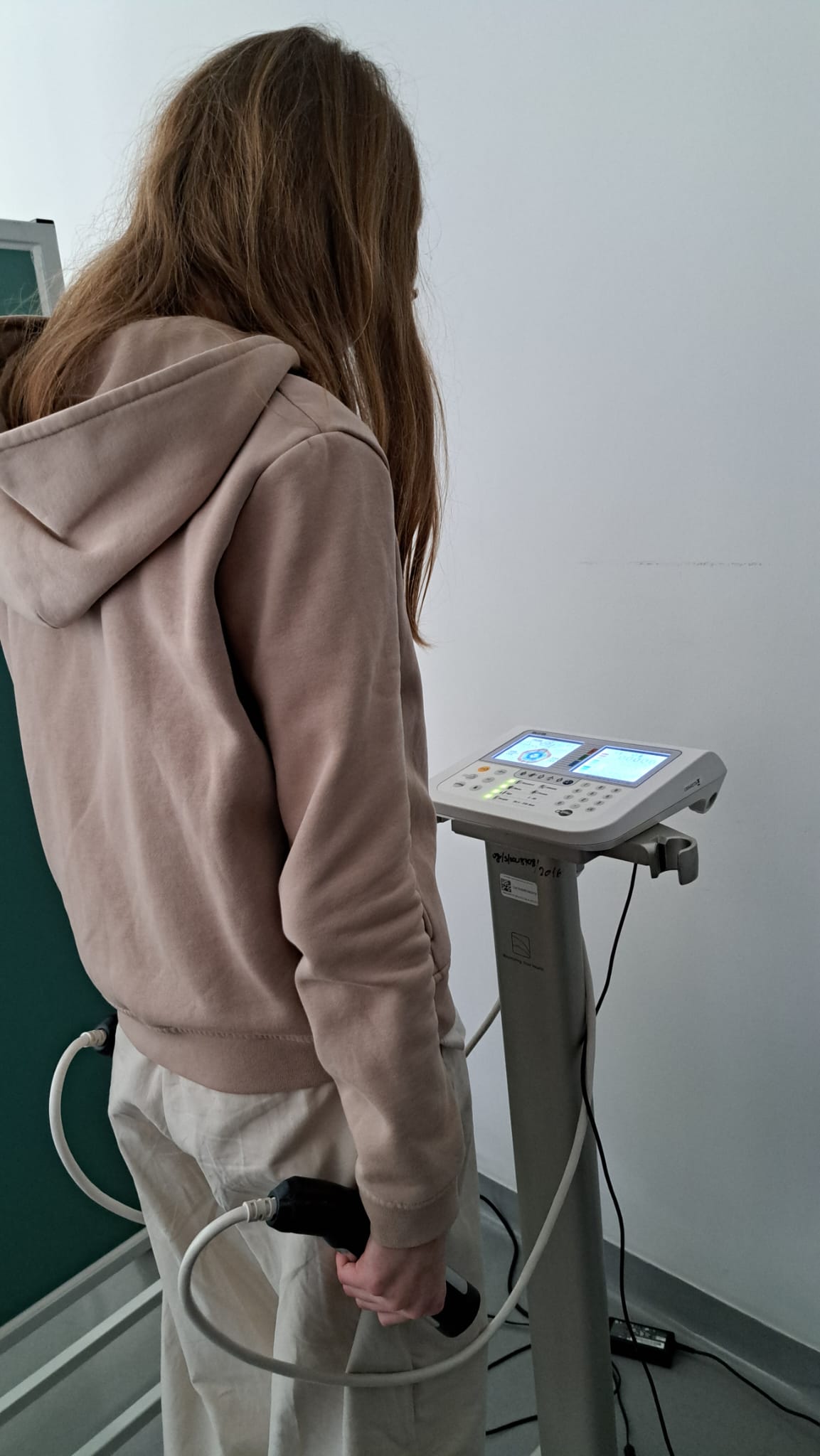 Obraz przedstawia osobę stojącą przy urządzeniu medycznym, trzymającą uchwyty podłączone do aparatu. Urządzenie ma panel sterowania z ekranami i przyciskami. Pomieszczenie jest proste, z jasnymi ścianami i podłogą oraz fragmentem tablicy po lewej.