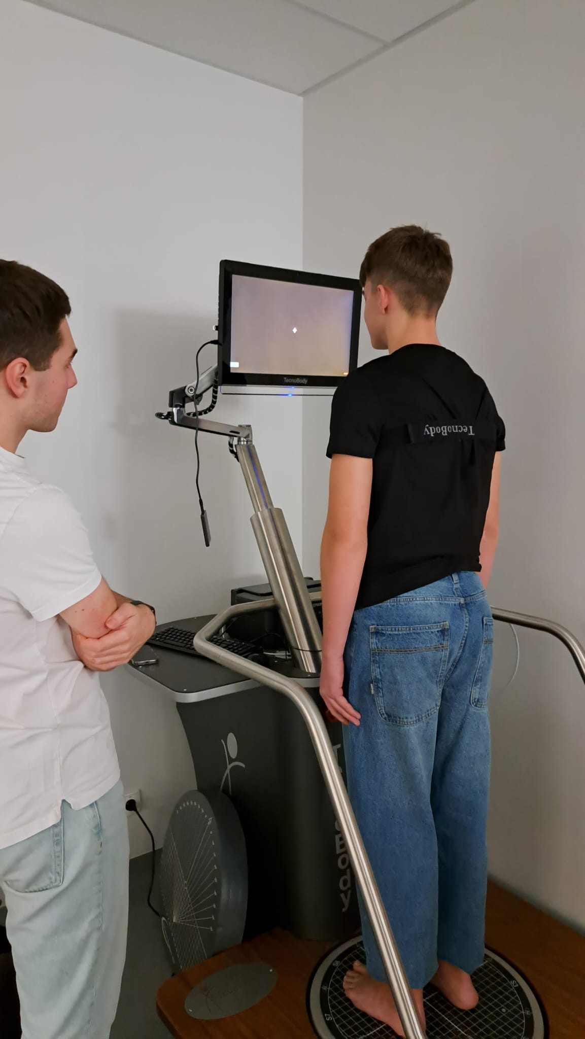 Obraz przedstawia dwóch młodych ludzi w pomieszczeniu klinicznym. Jeden stoi na platformie urządzenia do ćwiczeń lub równowagi, a drugi obserwuje go z boku. W tle widać białą ścianę i jasnoszarą podłogę.