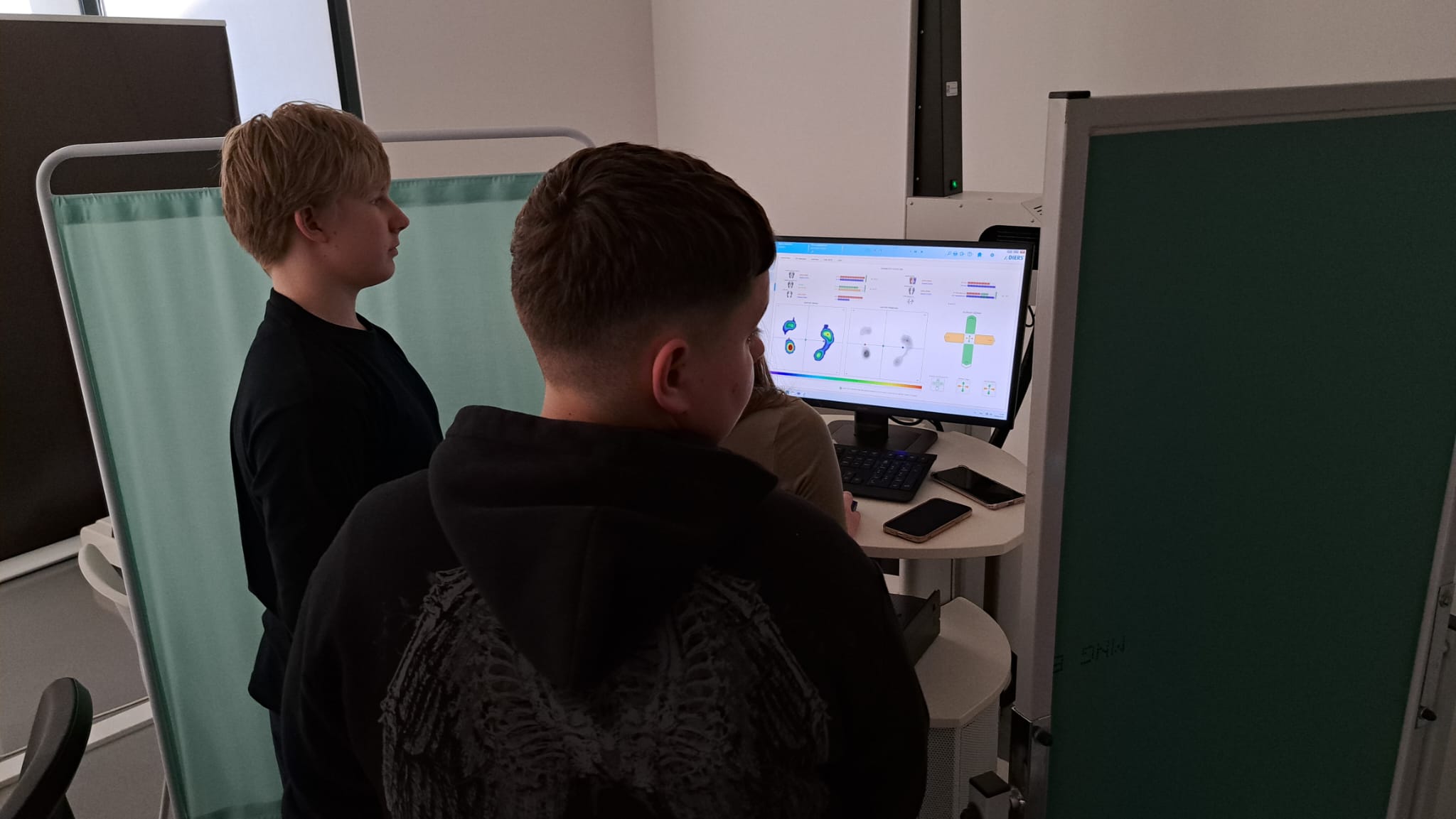 The image depicts two individuals standing in front of a computer monitor in a medical or laboratory setting. Both are viewing the screen attentively. The monitor displays various colorful graphs and charts, likely related to medical or scientific data. A light green privacy curtain is positioned on the left side, partially enclosing the space. A chair is visible in the lower left corner. The room is lit softly, suggesting an indoor environment with minimal natural light.