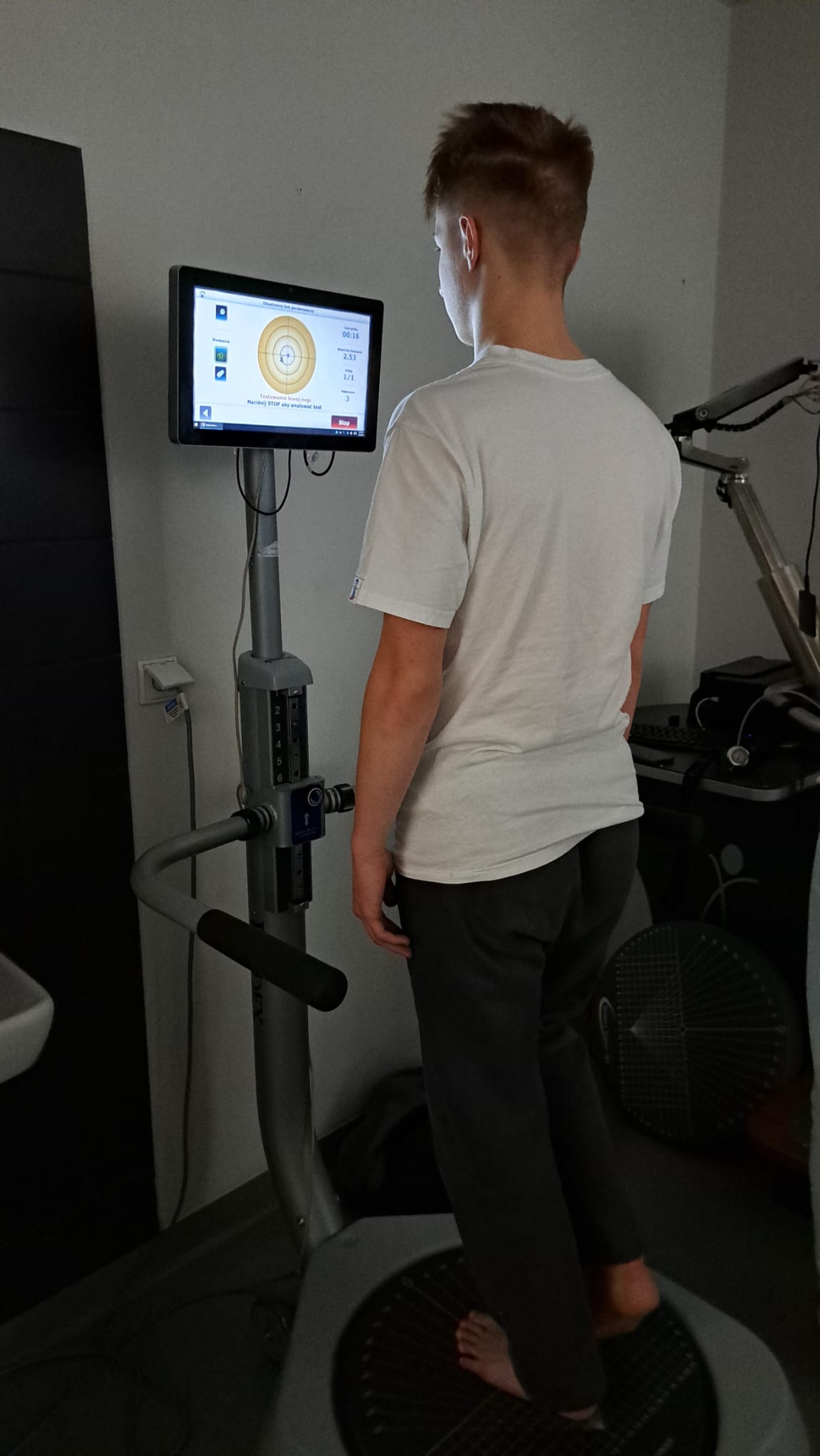 Obraz przedstawia osobę stojącą na platformie do treningu wibracyjnego w jasnym pomieszczeniu. Osoba patrzy na ekran zamontowany na platformie, a sama platforma ma teksturowaną powierzchnię, regulowane pokrętła i widoczne kable. Obok widać fragment innego urządzenia.