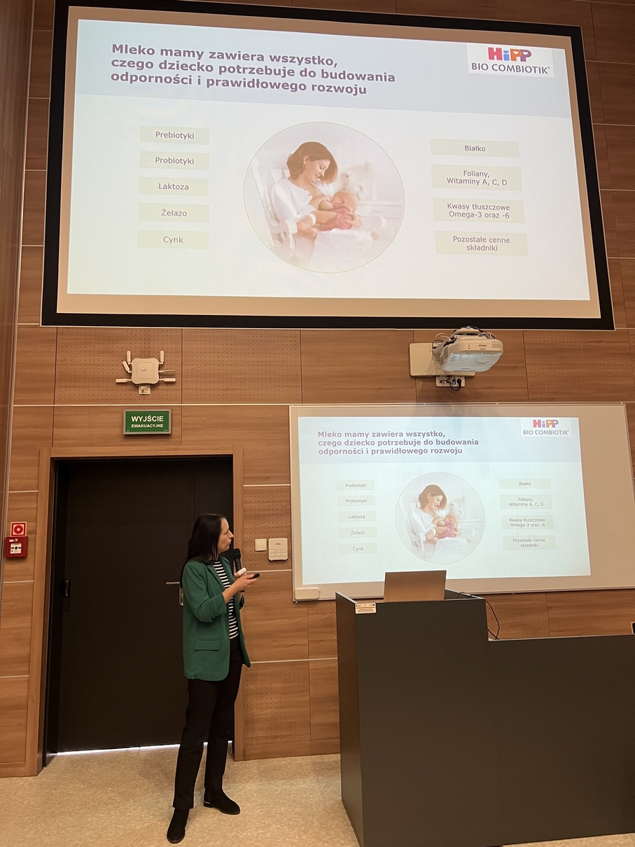 Osoba prowadzi prezentację w sali wykładowej, wskazując na slajd wyświetlany na dużym ekranie. Slajd zawiera grafikę z matką i dzieckiem oraz informacje dotyczące produktów HiPP Bio Combiotik. Wnętrze sali ma drewniane panele na ścianach, a w tle widoczne są drzwi z oznaczeniem wyjścia.