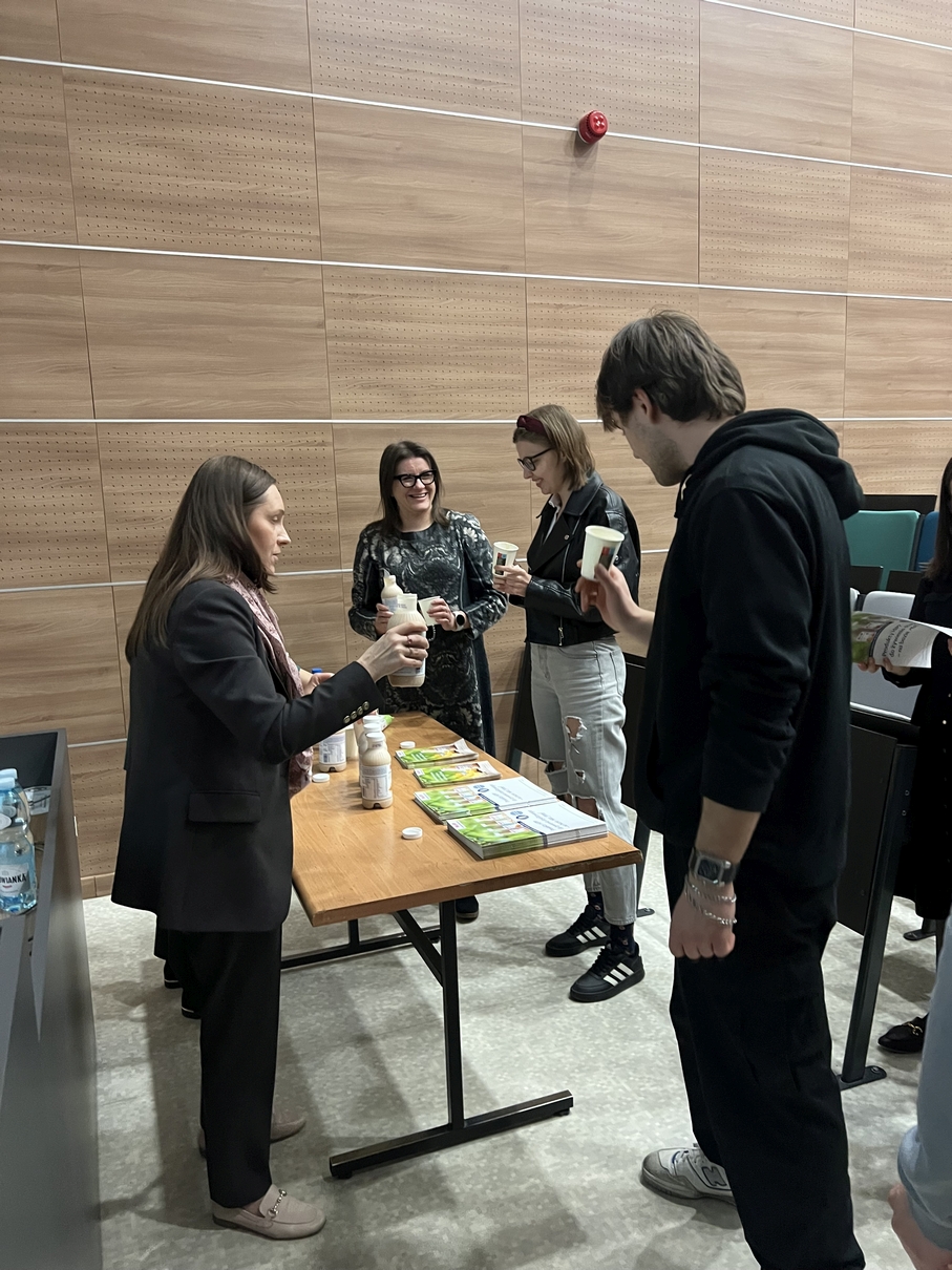 Grupa uczestników stoi przy stole w sali z jasnymi drewnianymi panelami na ścianach. Na stole znajdują się produkty oraz materiały informacyjne, które są omawiane podczas spotkania lub warsztatu. Uczestnicy przyglądają się prezentowanym materiałom, uczestnicząc w dyskusji lub demonstracji.