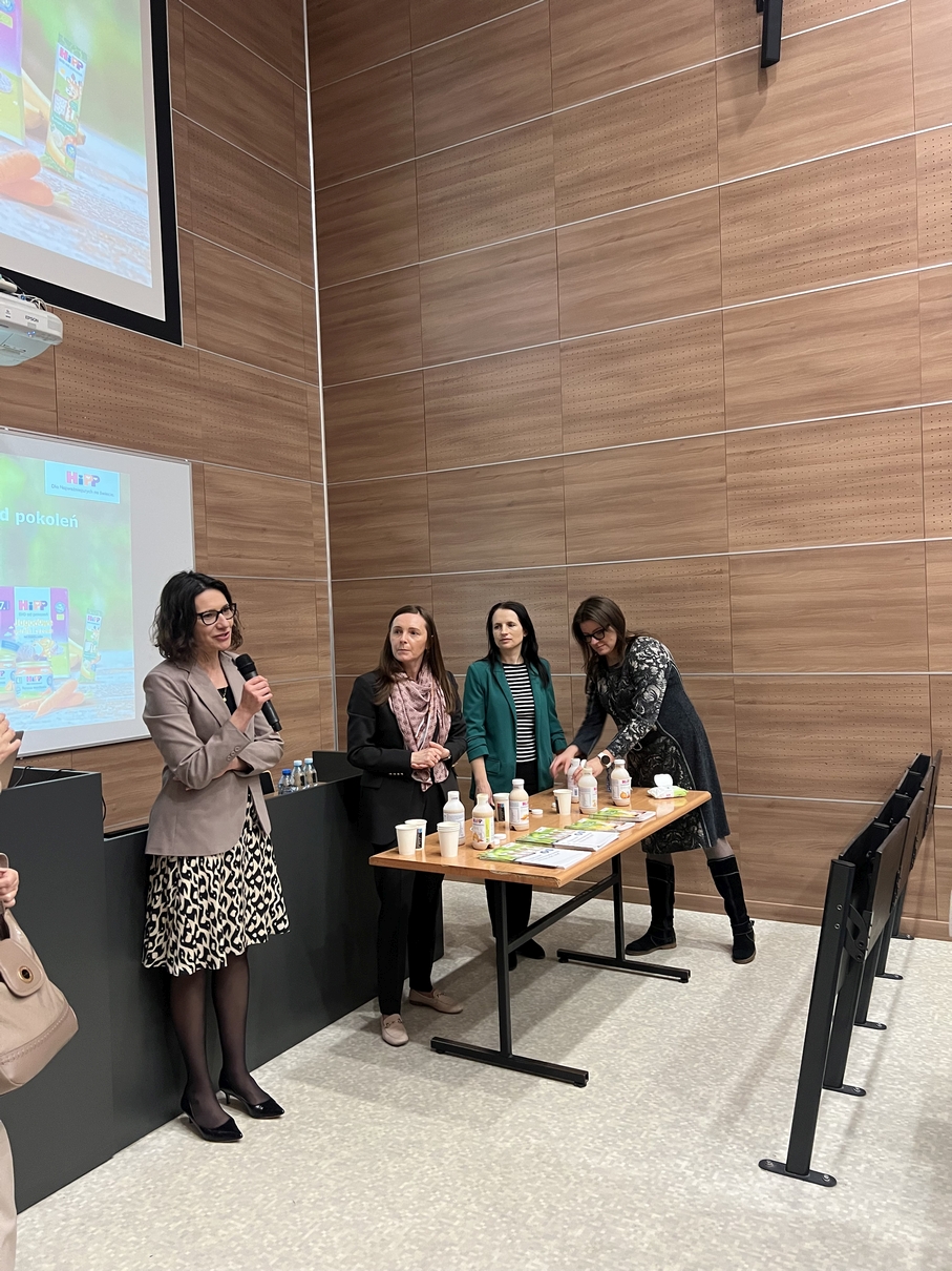 Cztery osoby stoją w sali wykładowej obok stołu z produktami oraz materiałami informacyjnymi. Jedna z osób przemawia do mikrofonu, prowadząc prezentację lub spotkanie. W tle widoczna jest duża projekcja z materiałami promocyjnymi oraz ściana z drewnianych paneli.