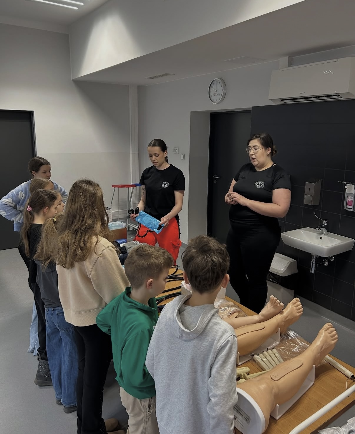 Dzieci zgromadzone wokół stołu uczestniczą w zajęciach edukacyjnych, obserwując demonstrację prowadzoną przez osoby prowadzące. Na stole znajdują się modele części ciała wykorzystywane do nauki. Sala ma jasne ściany, a w tle widoczny jest zegar oraz umywalka z dozownikiem mydła.
