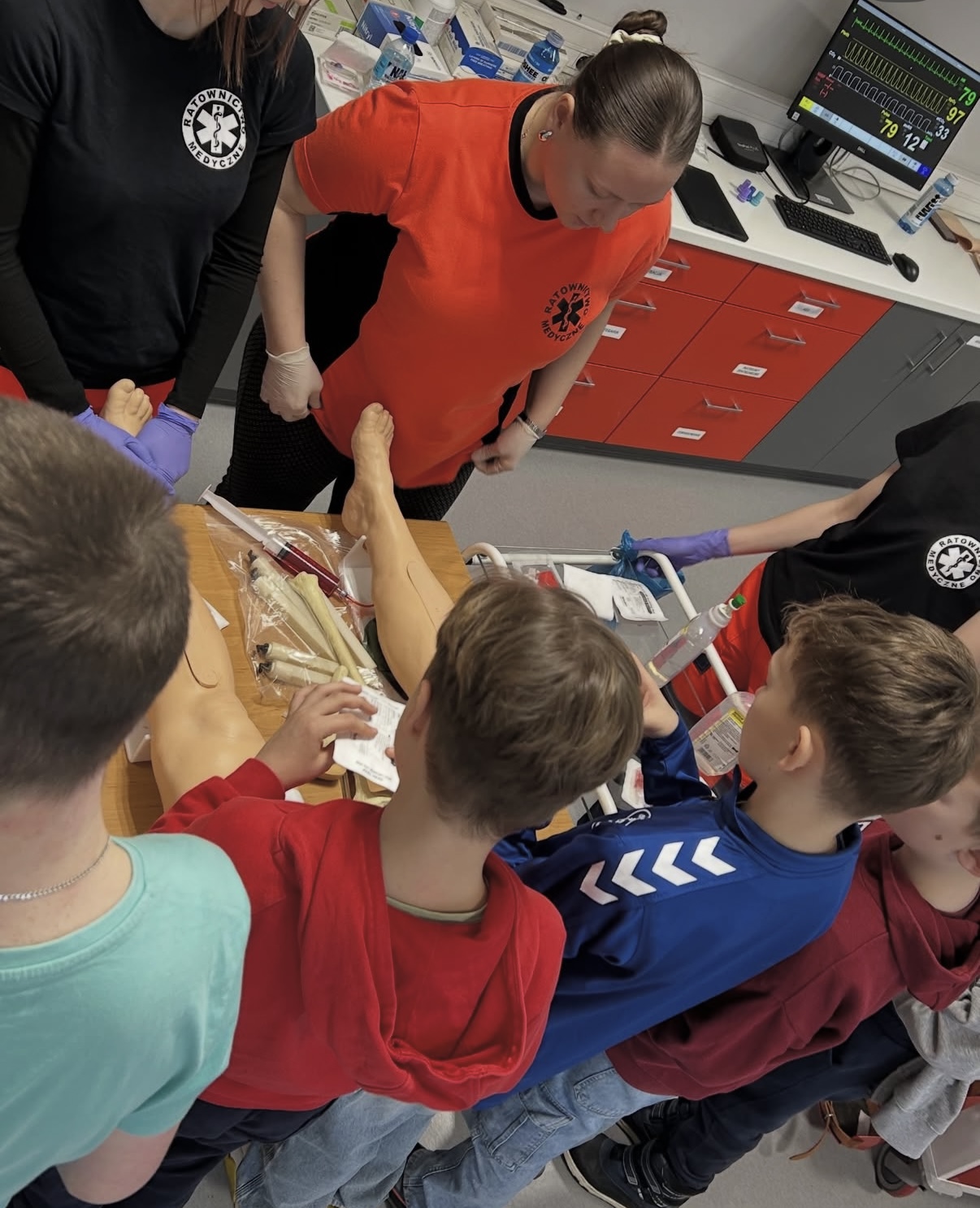 Grupa uczestników, w tym dzieci, zgromadzona jest wokół stołu z materiałami do nauki medycznej, takimi jak modele kończyn. Osoby prowadzące instruują i nadzorują ćwiczenia praktyczne. W tle widoczne są szafki, sprzęt medyczny oraz monitor z parametrami, co wskazuje na salę szkoleniową o charakterze medycznym.
