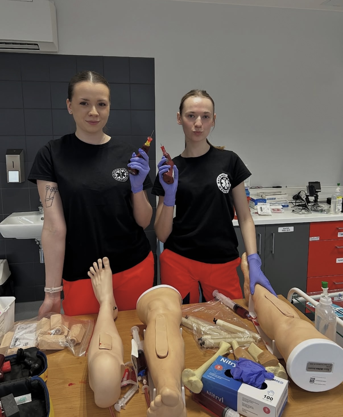 Dwie osoby stoją w sali szkoleniowej o charakterze medycznym, prezentując sprzęt wykorzystywany do nauki procedur. Na stole znajdują się modele części ciała, strzykawki oraz inne materiały szkoleniowe. W tle widoczne są półki z wyposażeniem, zlew oraz ściana wykończona ciemnymi płytkami.